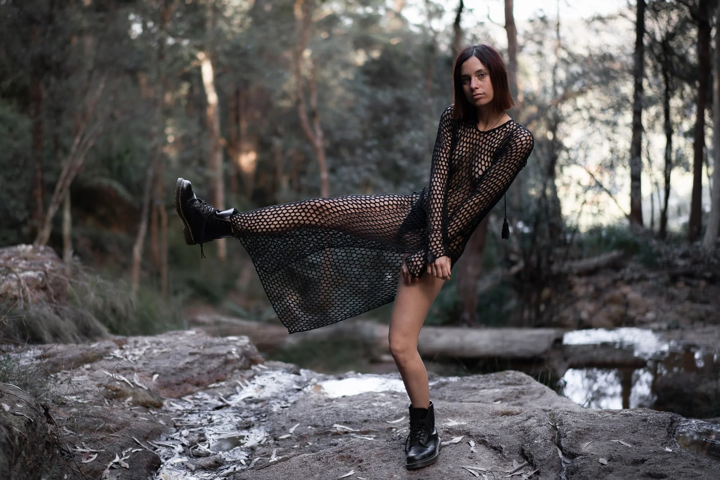 A young woman in a black fishnet dress and black combat boots standing on a rocky forest path, with one leg raised and her foot extended across her body, in a wooded outdoor setting with trees and a small creek in the background.