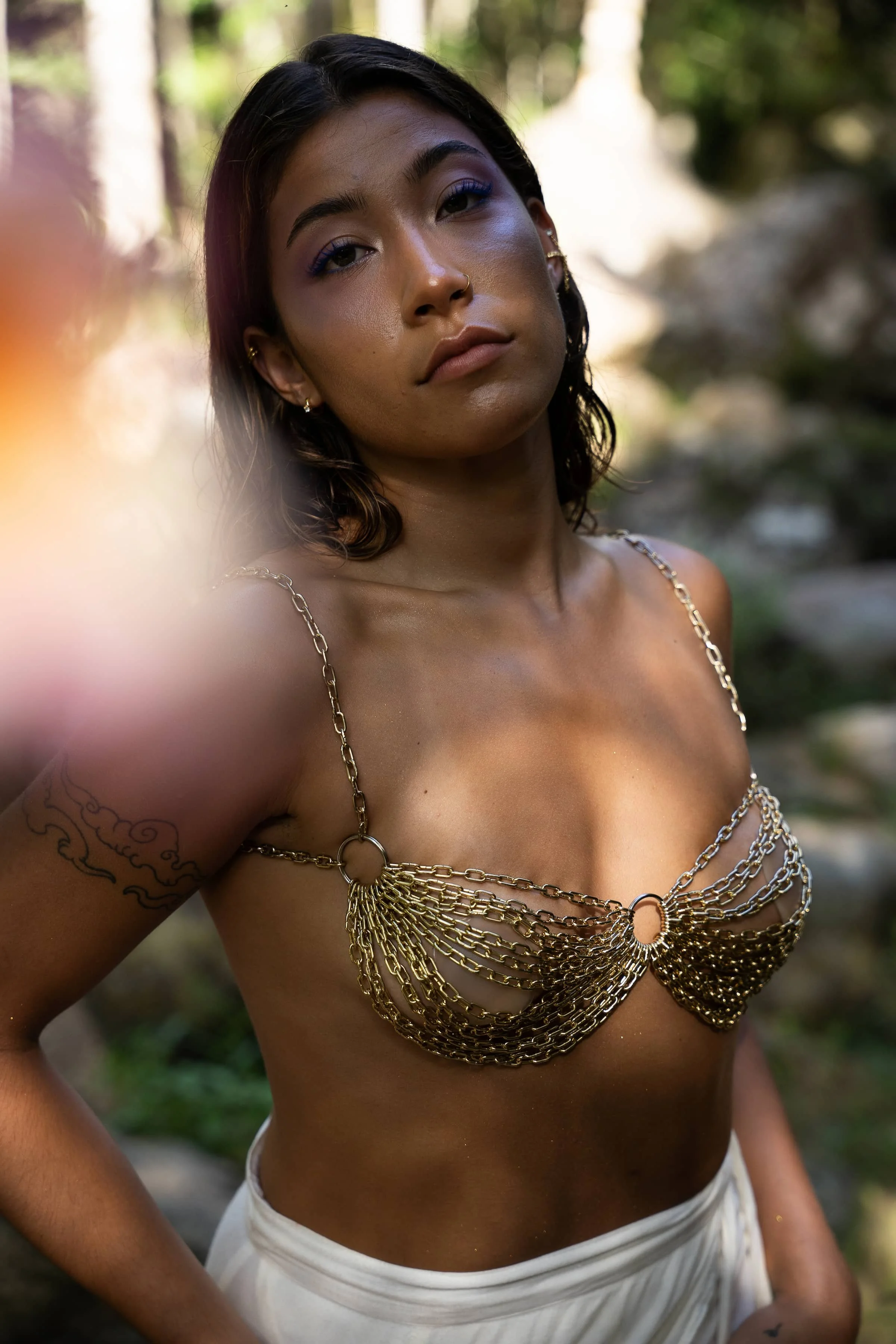 A woman with shoulder-length dark hair and a tattoo on her arm stands outdoors. She wears a decorative gold chain harness top and a white skirt, with a serious expression on her face.