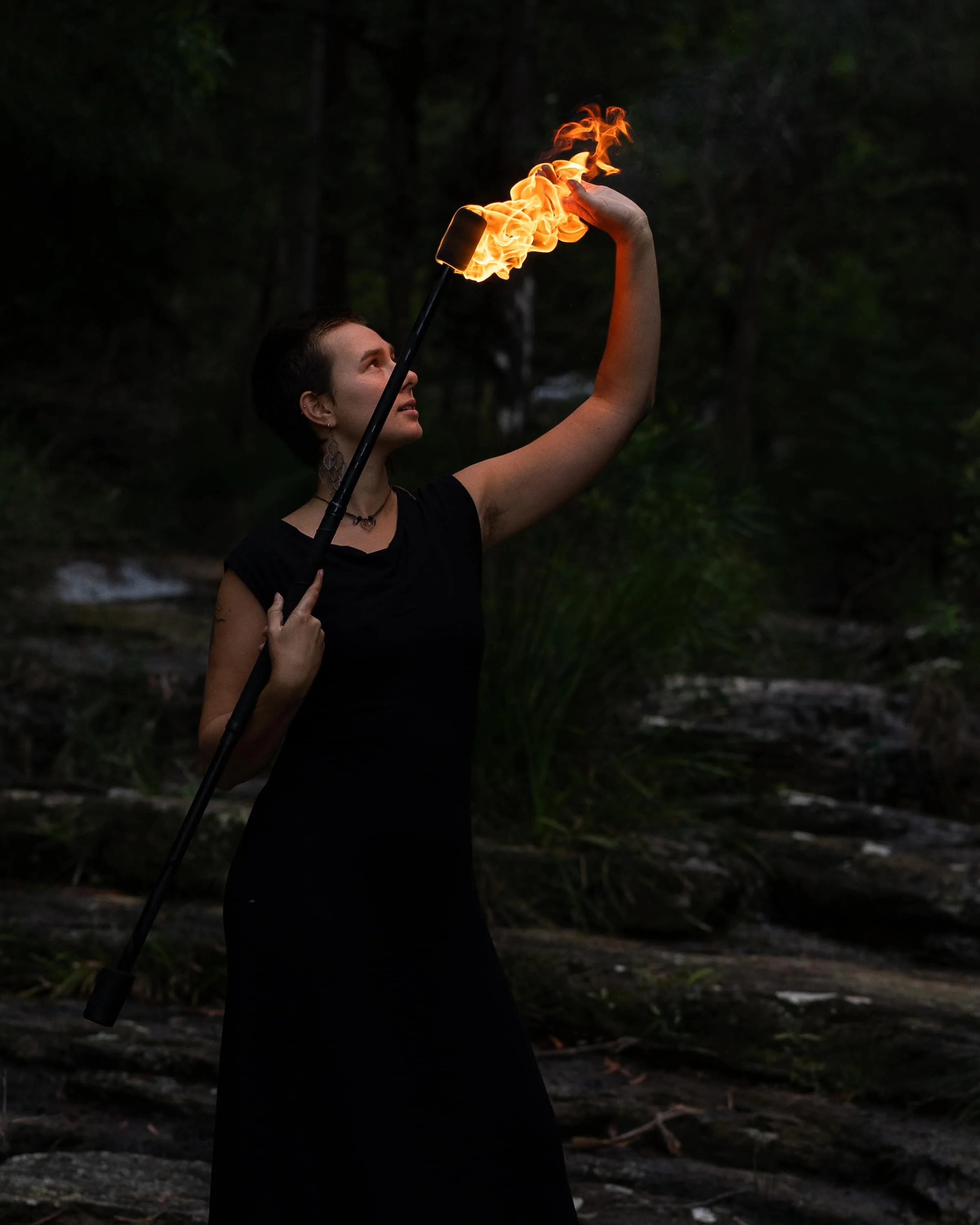A woman in a black dress performs a fire juggling act outdoors at dusk, holding a flaming torch in one hand and a torch with fire in the other, with a forest background.