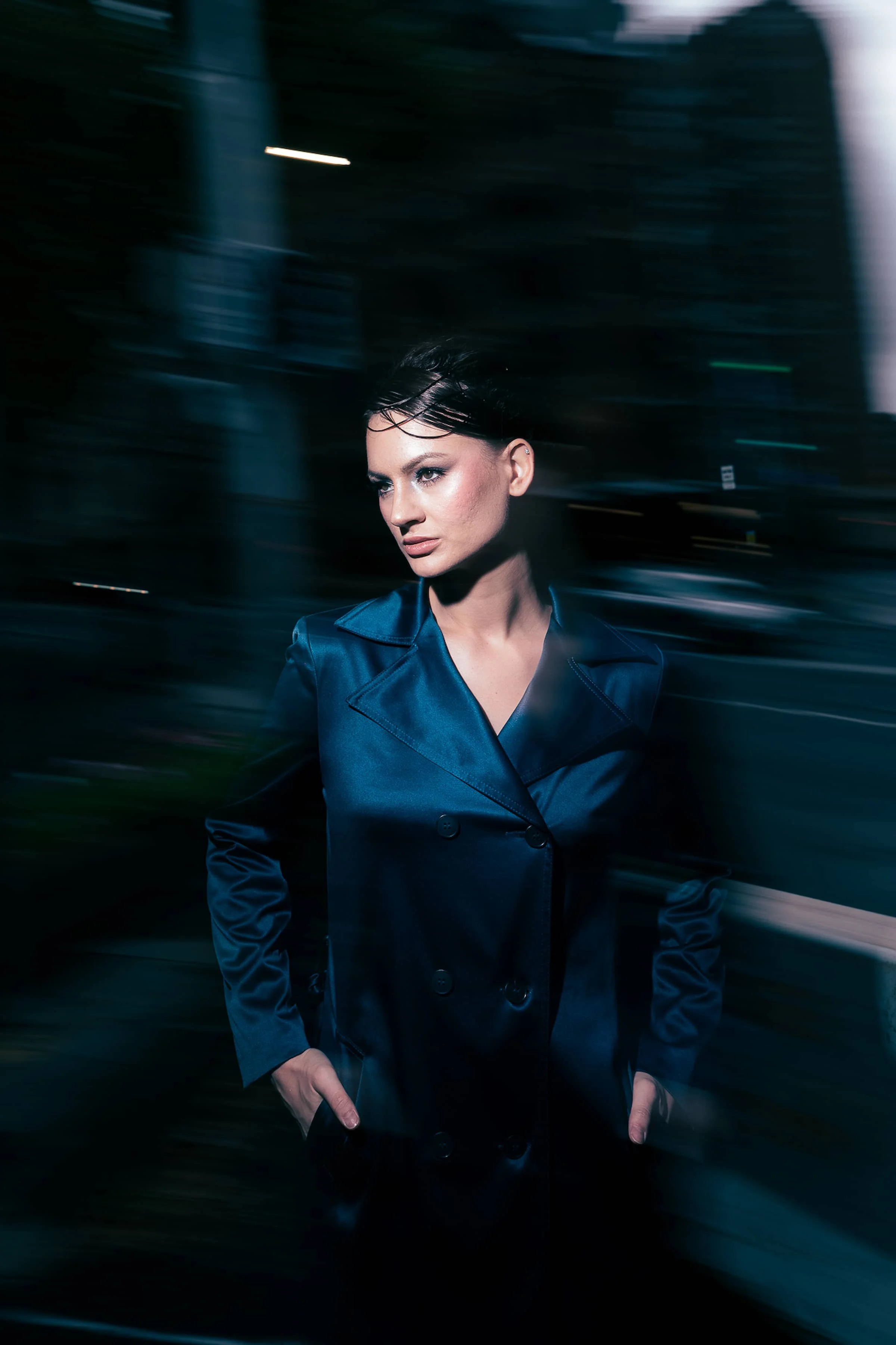 A woman in a shiny blue coat stands with her hands in her pockets against a blurred city background at night.