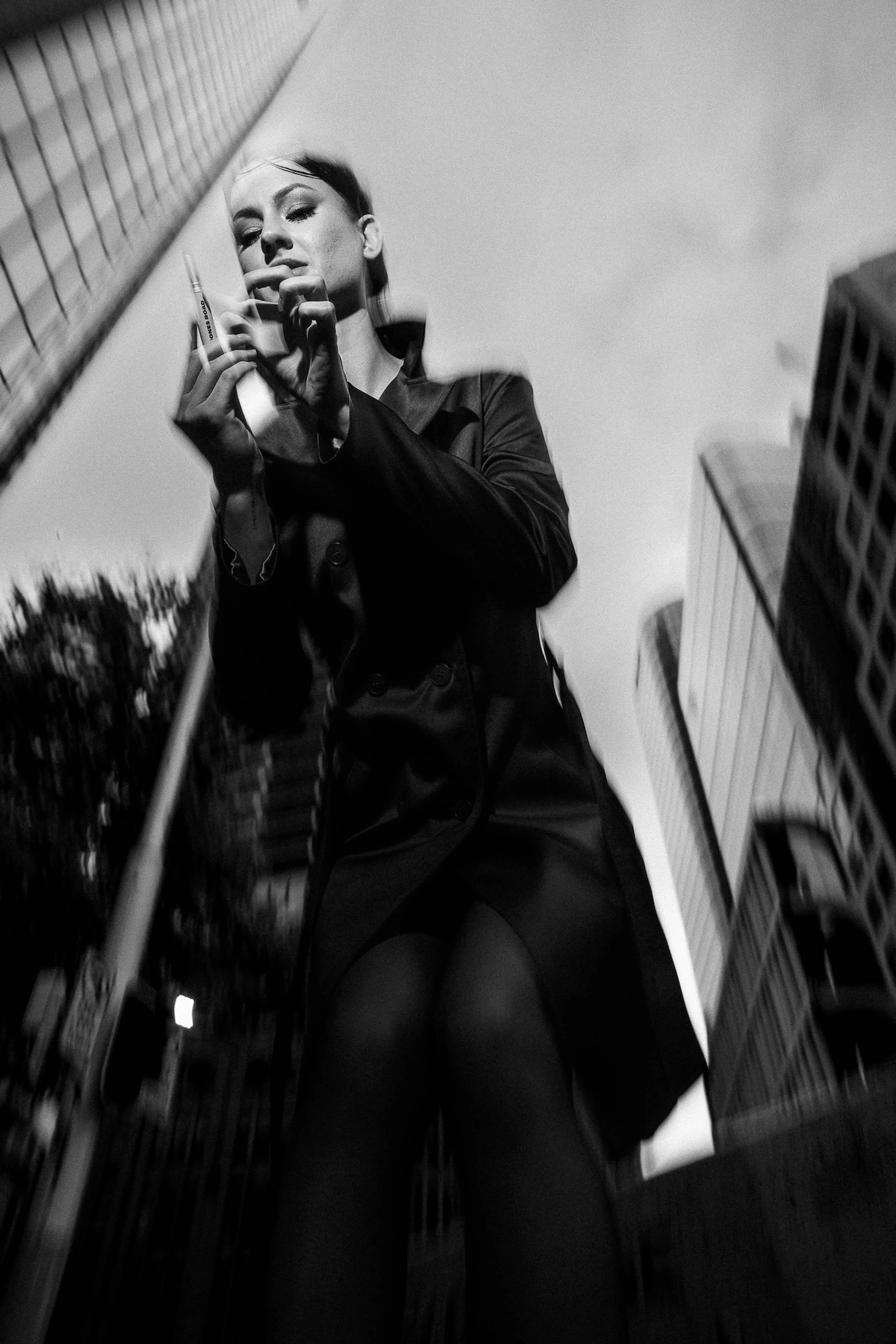 A woman dressed in dark clothing stands among tall buildings, looking down at a reflective surface she is holding, with her reflection visible in it.