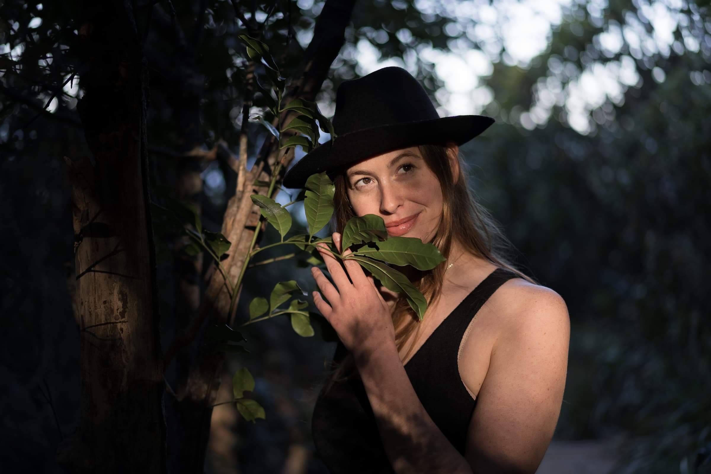 A woman with reddish-brown hair is wearing a black hat and black tank top, smiling softly while holding green leaves near her face in an outdoor setting with trees and natural light.