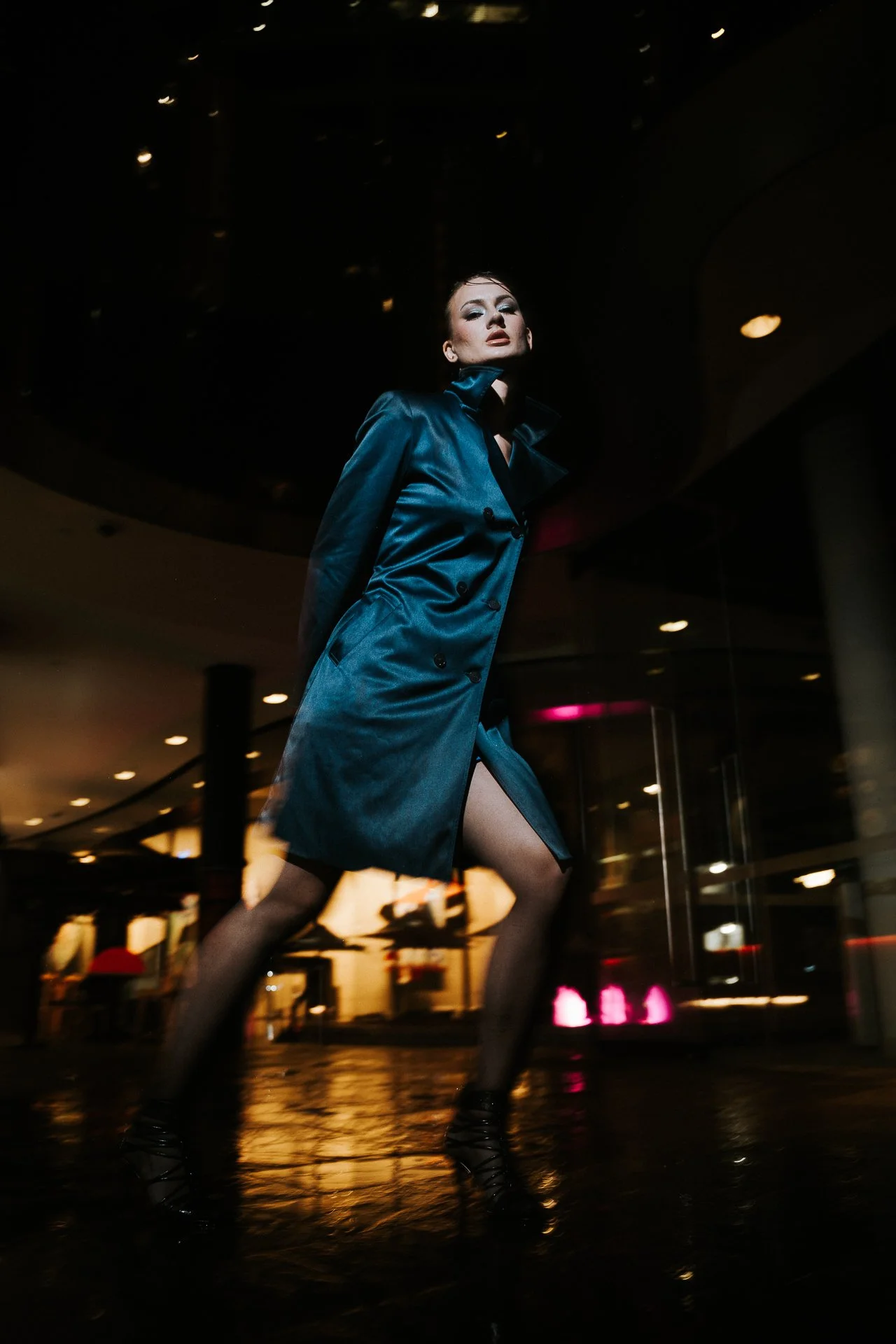 A woman in a shiny blue trench coat and high heels is posing confidently in a dark, modern indoor space with dramatic lighting.