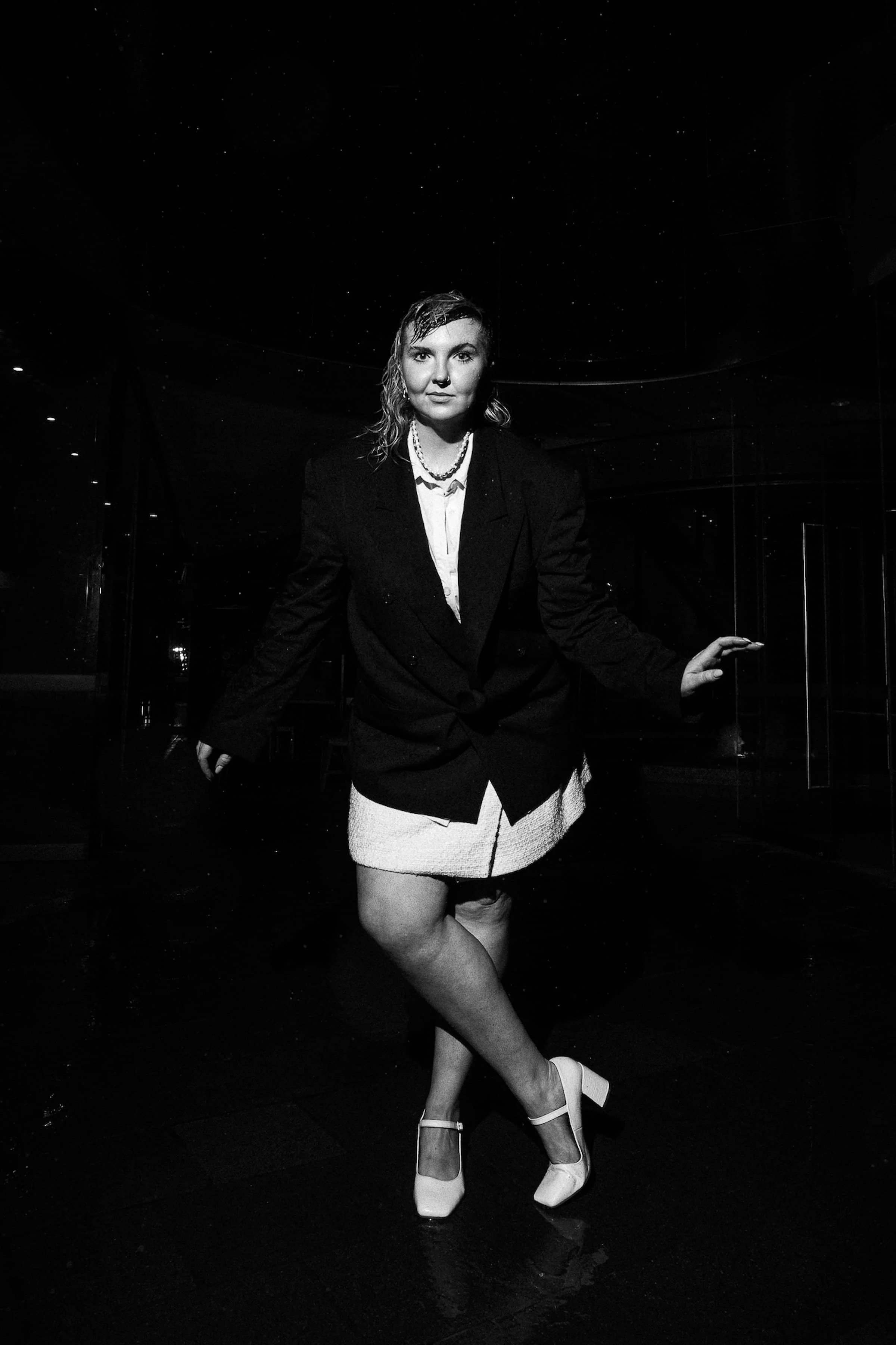 Black and white photo of a woman wearing a blazer, a skirt, and high heels, standing with one leg crossed over the other and one hand resting on a surface, in a dimly lit indoor space.
