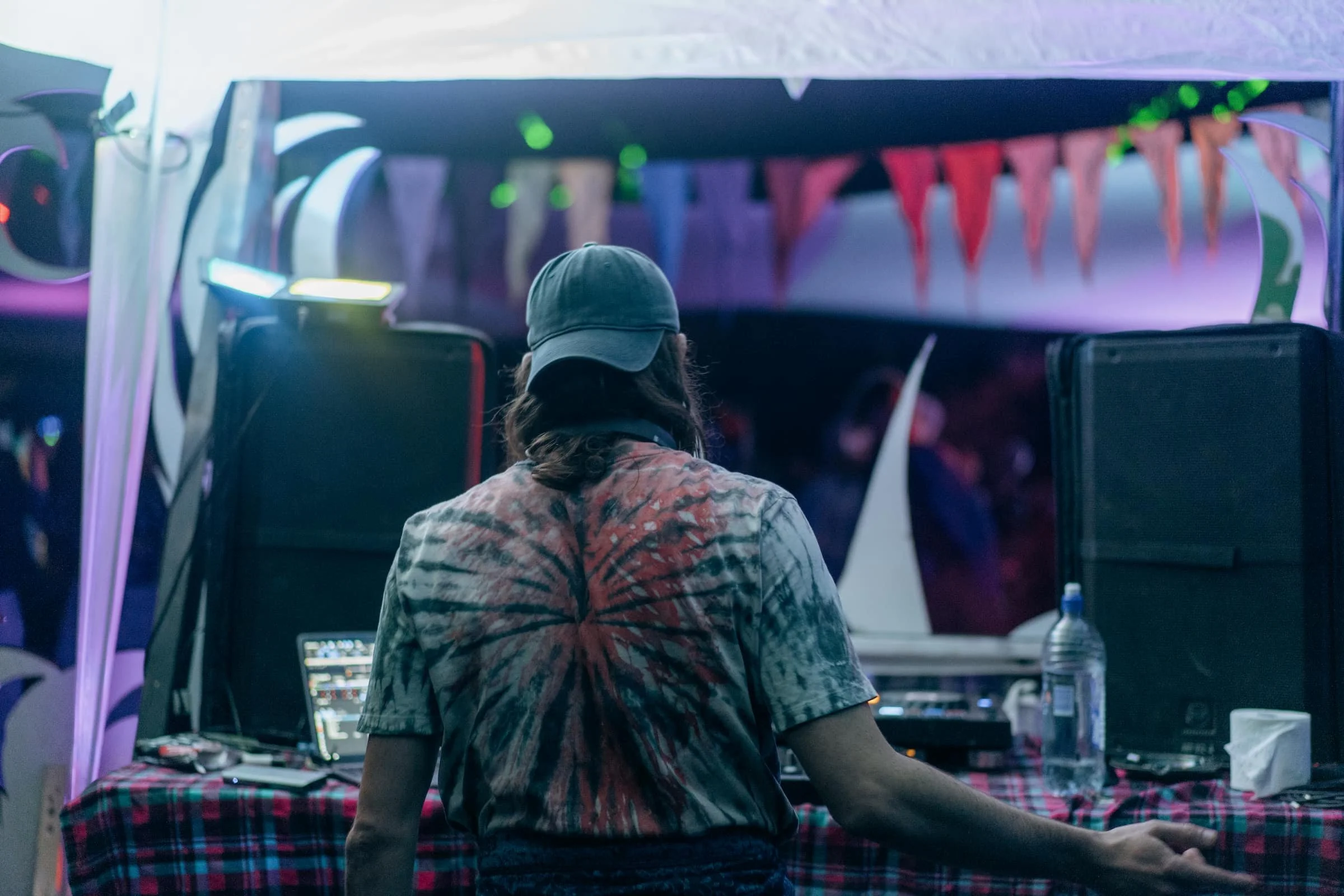 Person DJing at a party or event, with DJ equipment and speakers on a table, colorful decorations, and illuminated background.