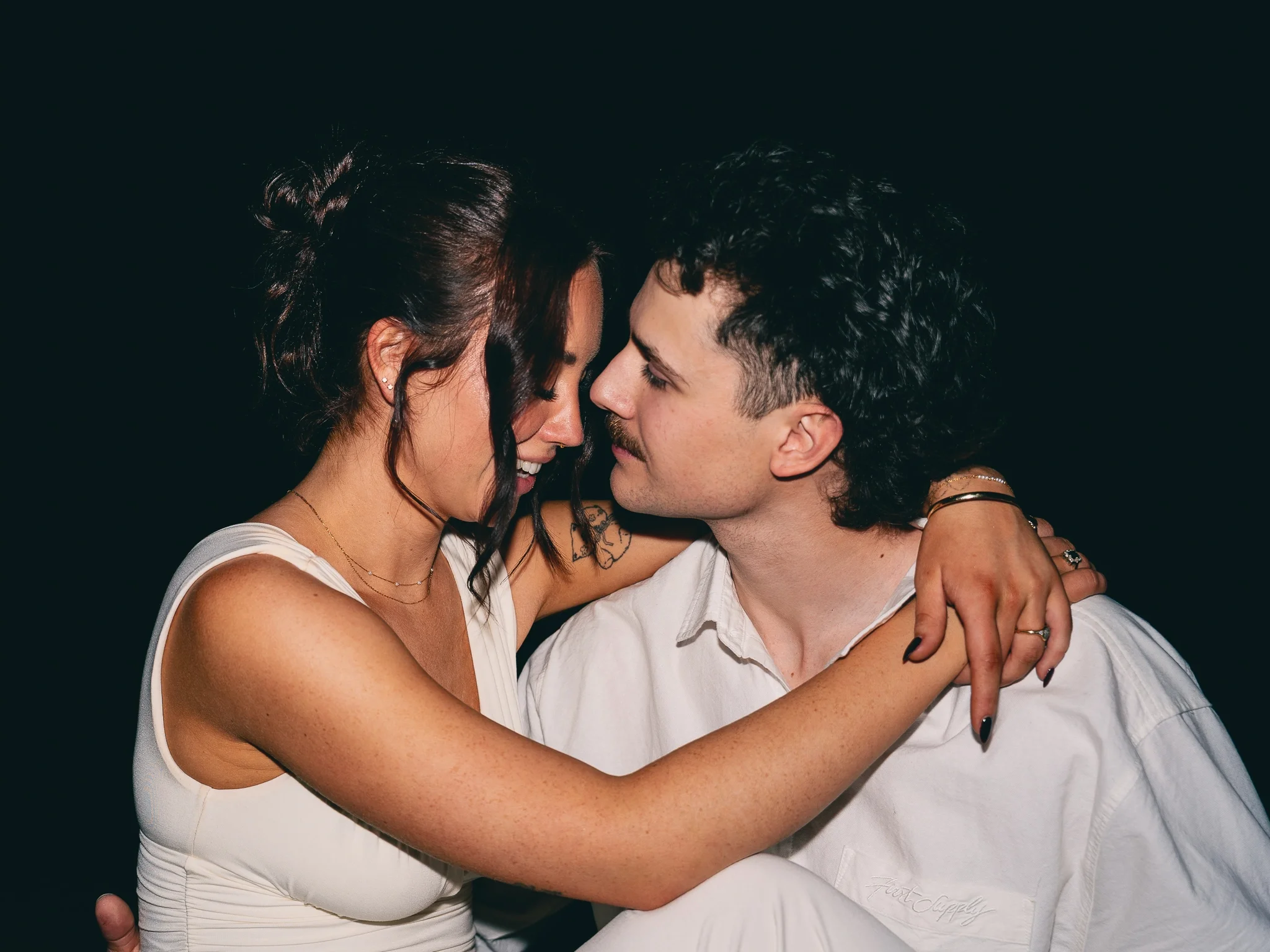 A young couple with light skin and dark hair sharing an intimate moment, noses touching, and smiling with their eyes closed, against a plain dark background.