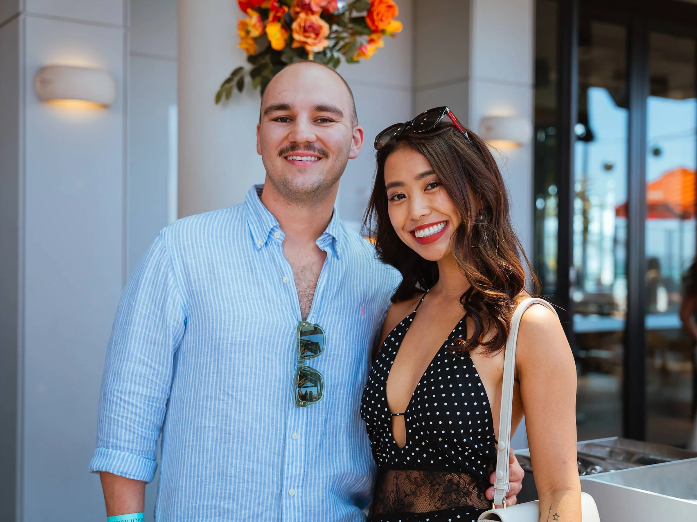 A smiling man and woman posing together outdoors near a restaurant or cafe. The man is wearing a light blue, striped button-up shirt with sunglasses hanging on the front. The woman has shoulder-length wavy hair, wears a black polka-dot dress, red lip
