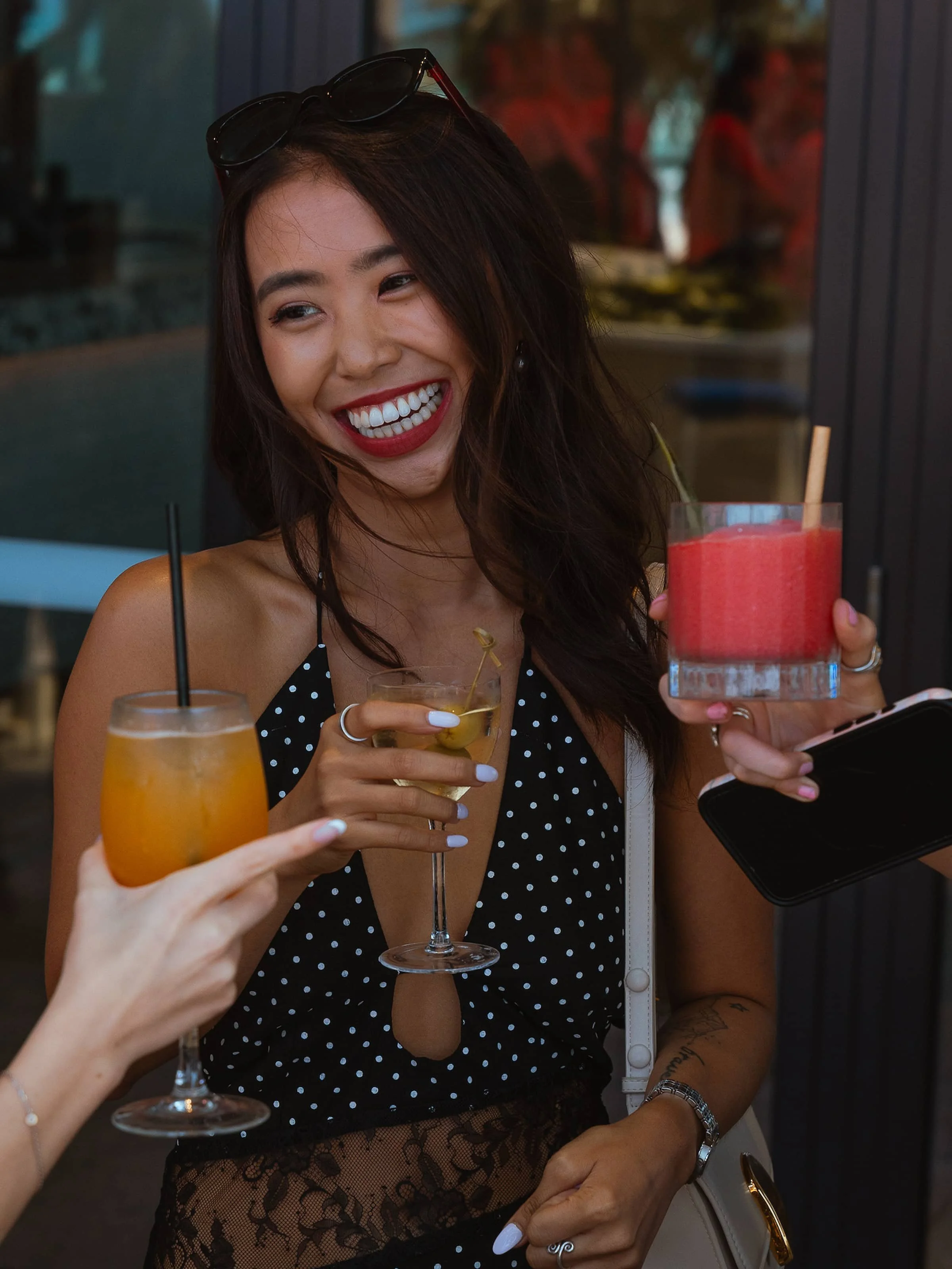 A woman with long dark hair, wearing a black and white polka dot top, smiling widely while holding a pink cocktail. Two other people, only their hands visible, are holding different drinks and appear to be toast/cheers with her. The background sugges