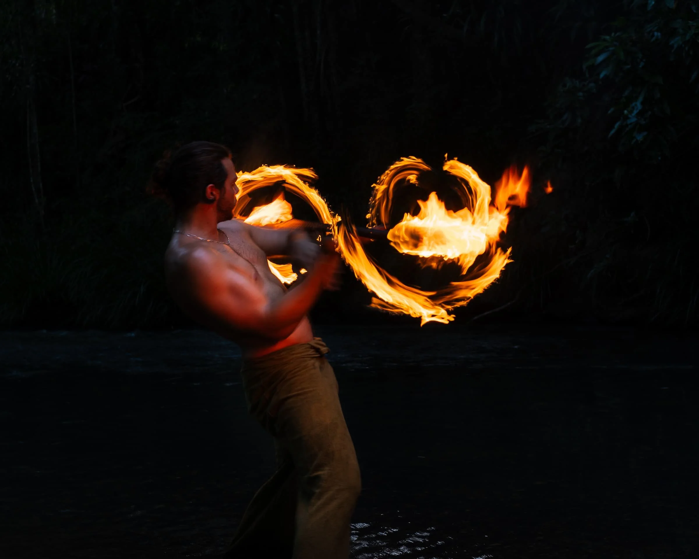 A shirtless man with long hair tied back and wearing brown pants is performing fire poi juggling in a dark outdoor setting.
