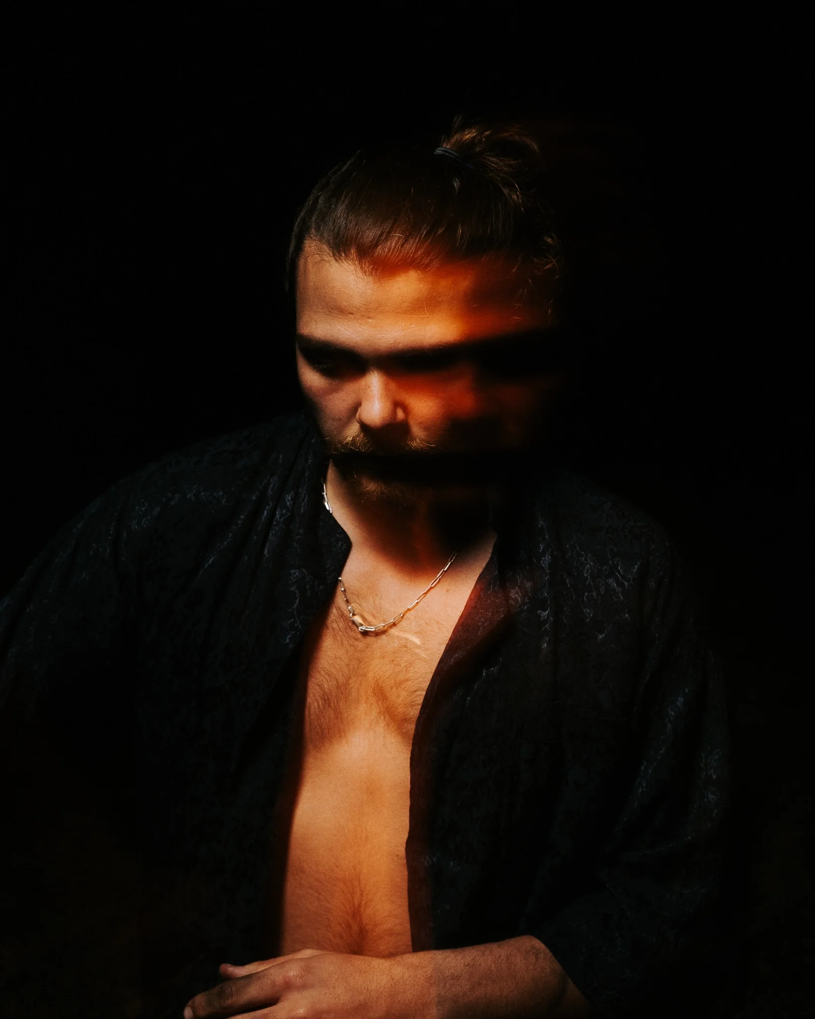 A person with a partially covered face, wearing a dark jacket, with a chain necklace visible, in a dark background.