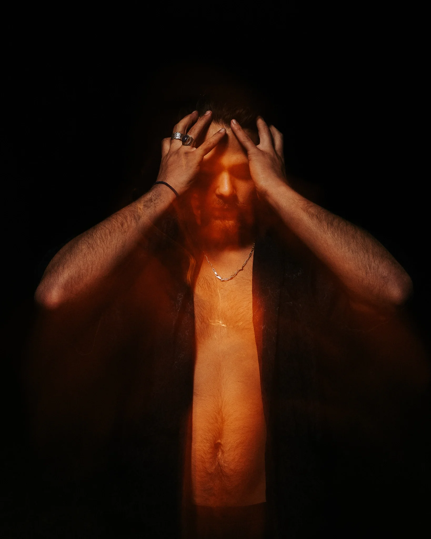 A person with a dark beard and short brown hair holds their head with both hands, fingers pressing against their forehead, in front of a black background with a fiery glow in the center.