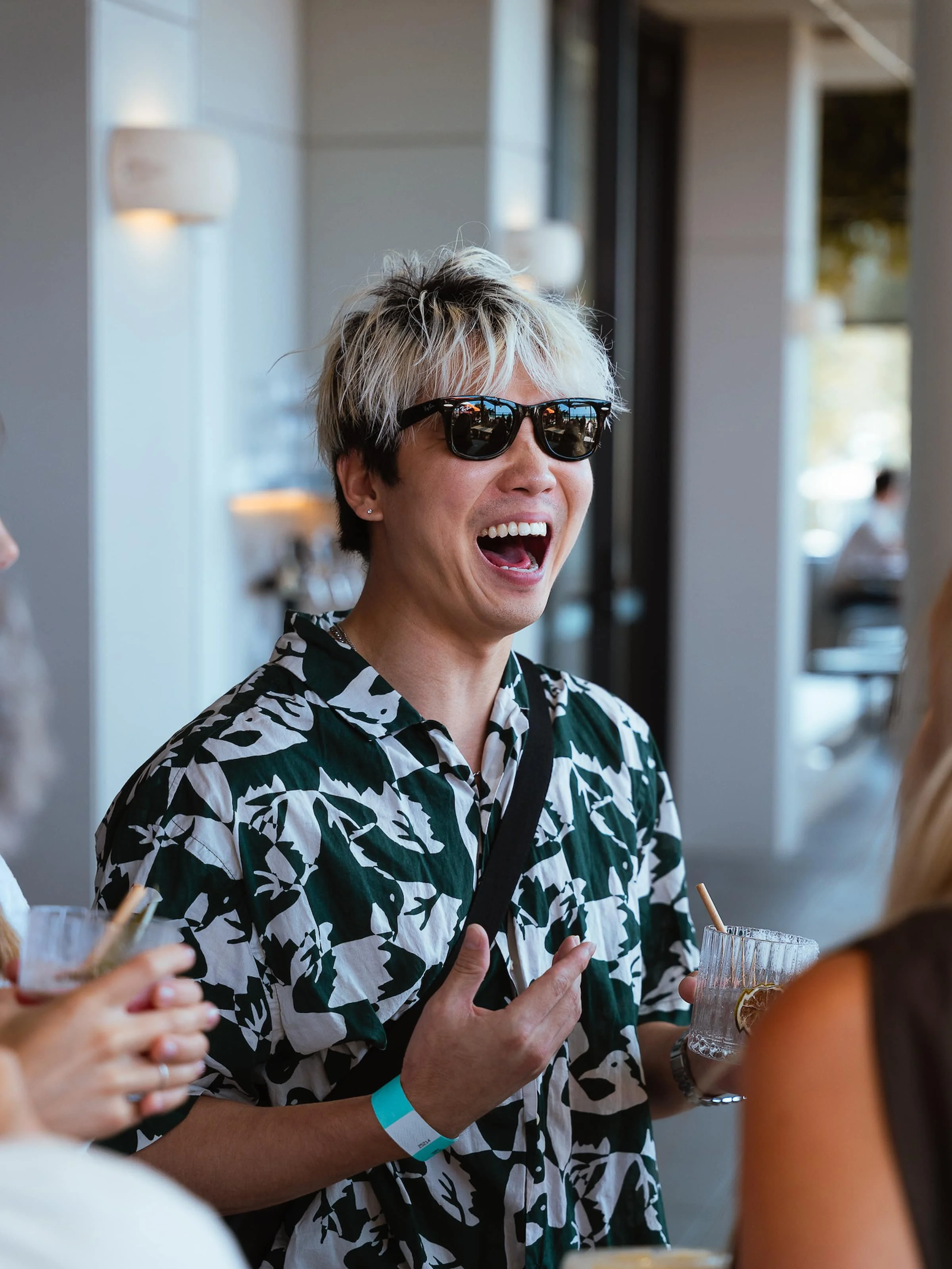 A young man with blond and black hair, wearing sunglasses and a black and white patterned shirt, is laughing and talking with others at a social gathering, holding a glass with a drink.