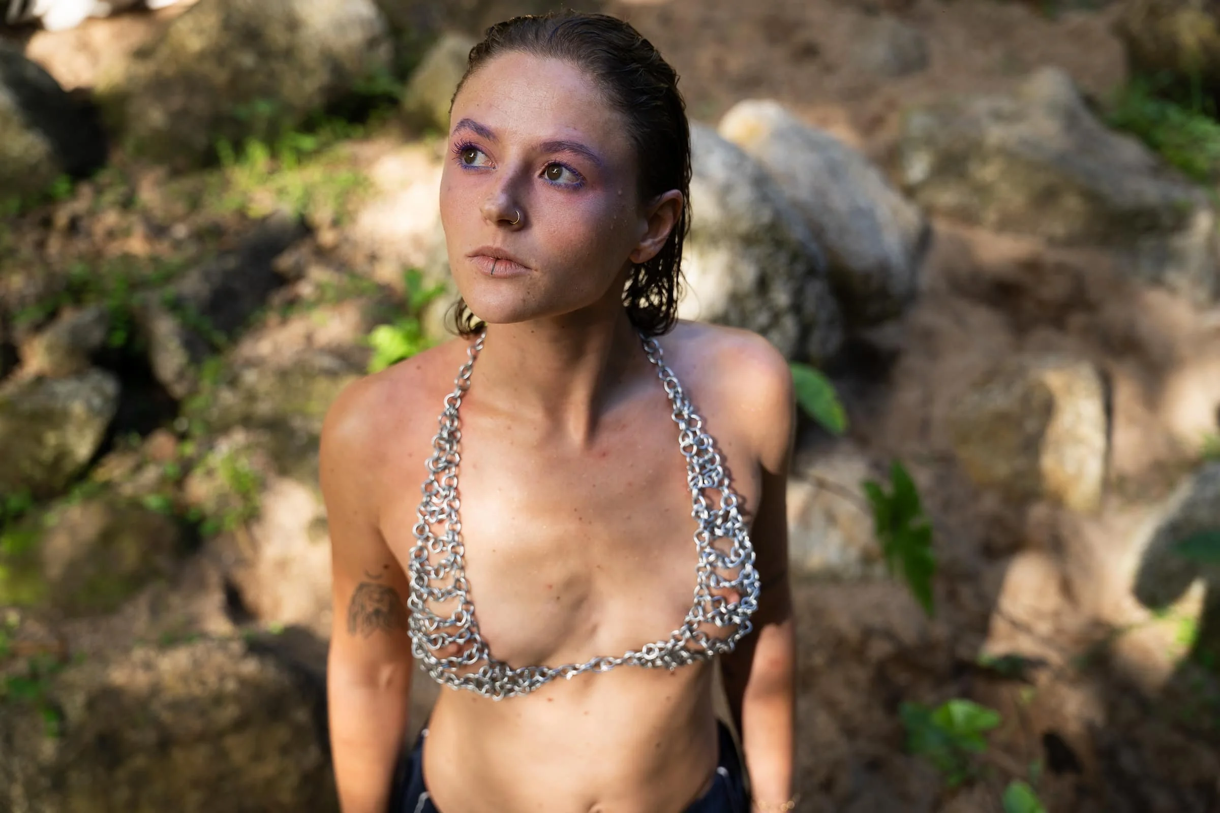 Young woman with wet hair and minimal makeup, wearing a chunky silver chain necklace, posing outdoors among rocks and greenery.