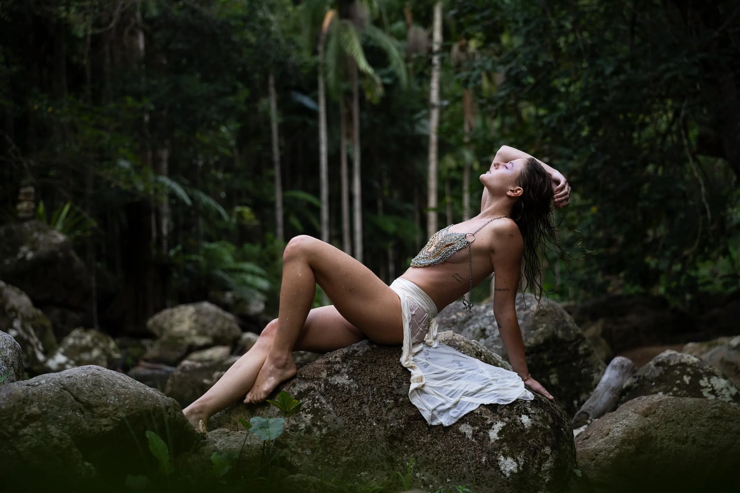 A woman in a white skirt decorated with chains, sitting on a rock in a lush forest, with her head tilted back and eyes closed.