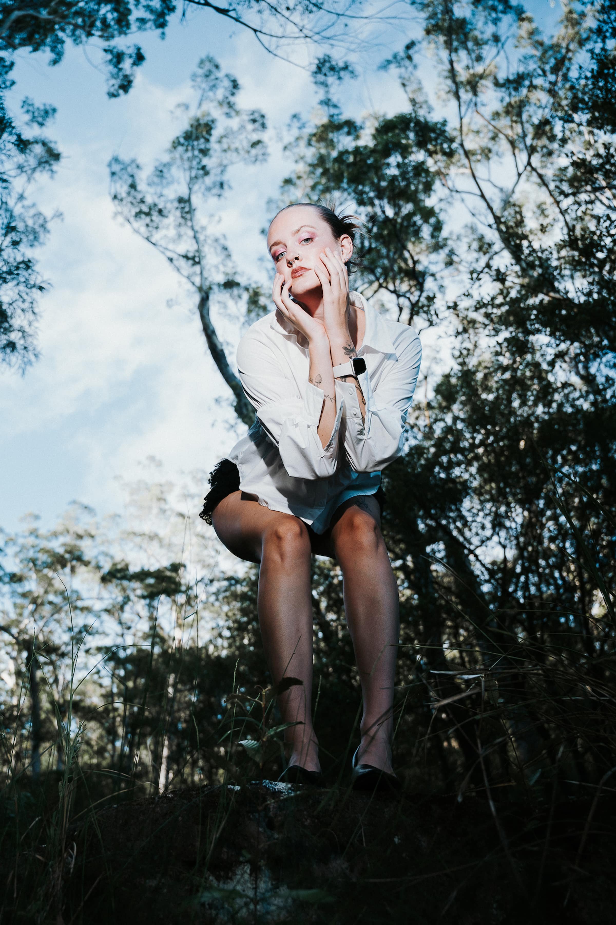 A woman is crouching outdoors among trees, with her hands on her cheeks and an expressive pose. She is wearing a white shirt, black shorts, and a smartwatch, with a blue sky visible through the trees.