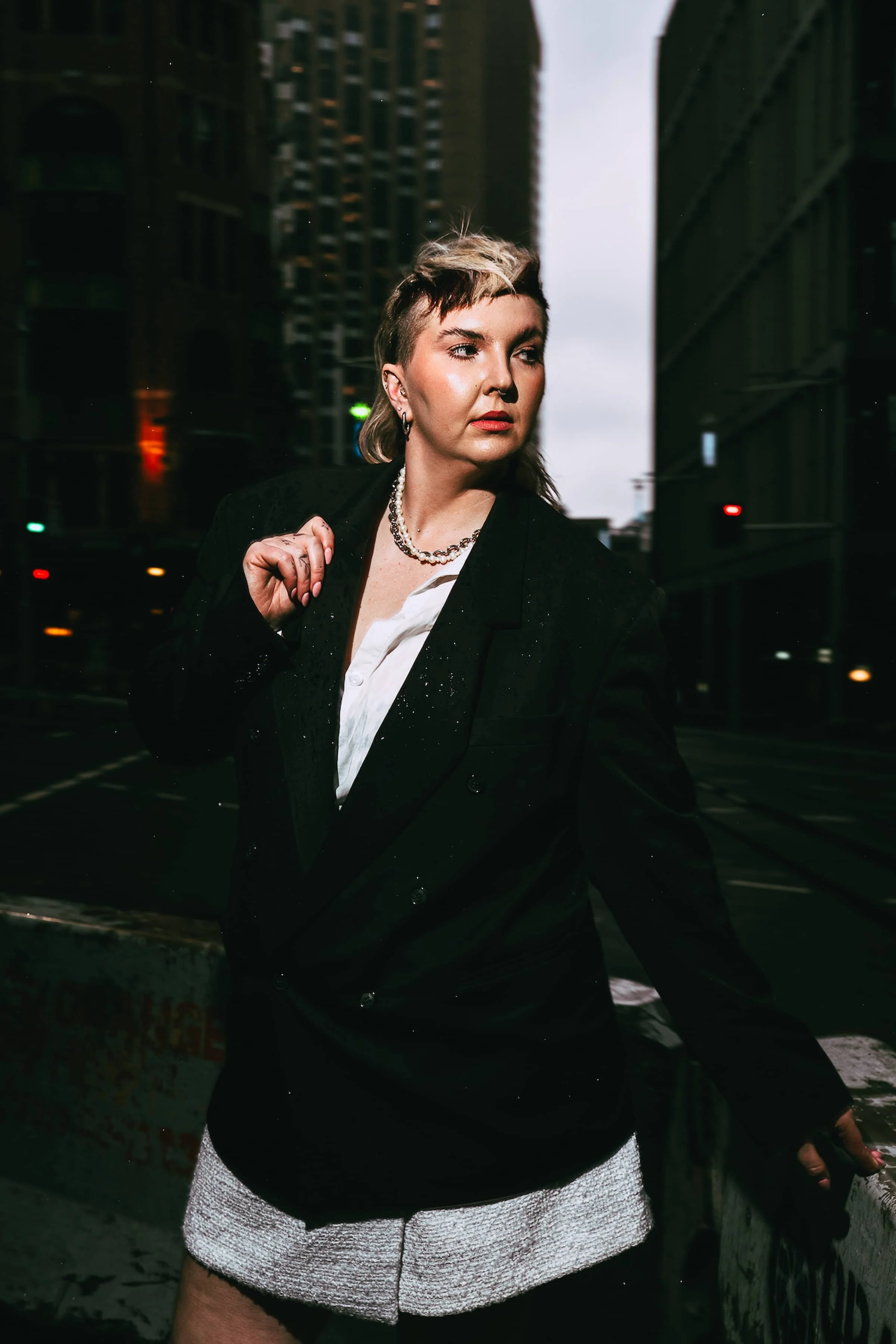 A woman in a black blazer and white top standing outdoors at night in an urban environment with tall buildings in the background.