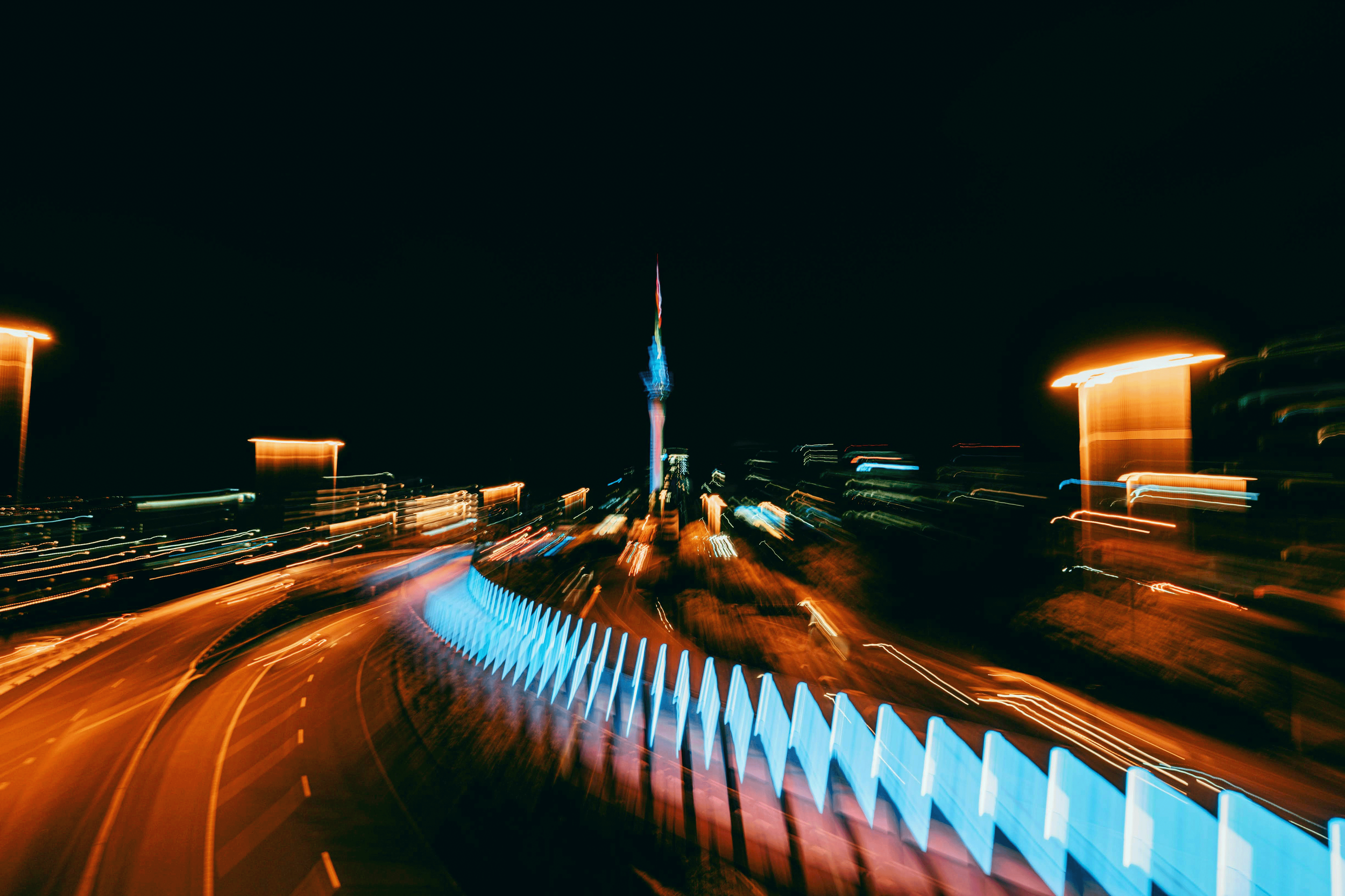 Nighttime cityscape with blurred streaks of moving traffic and colorful lights, featuring a tall illuminated tower in the distance.