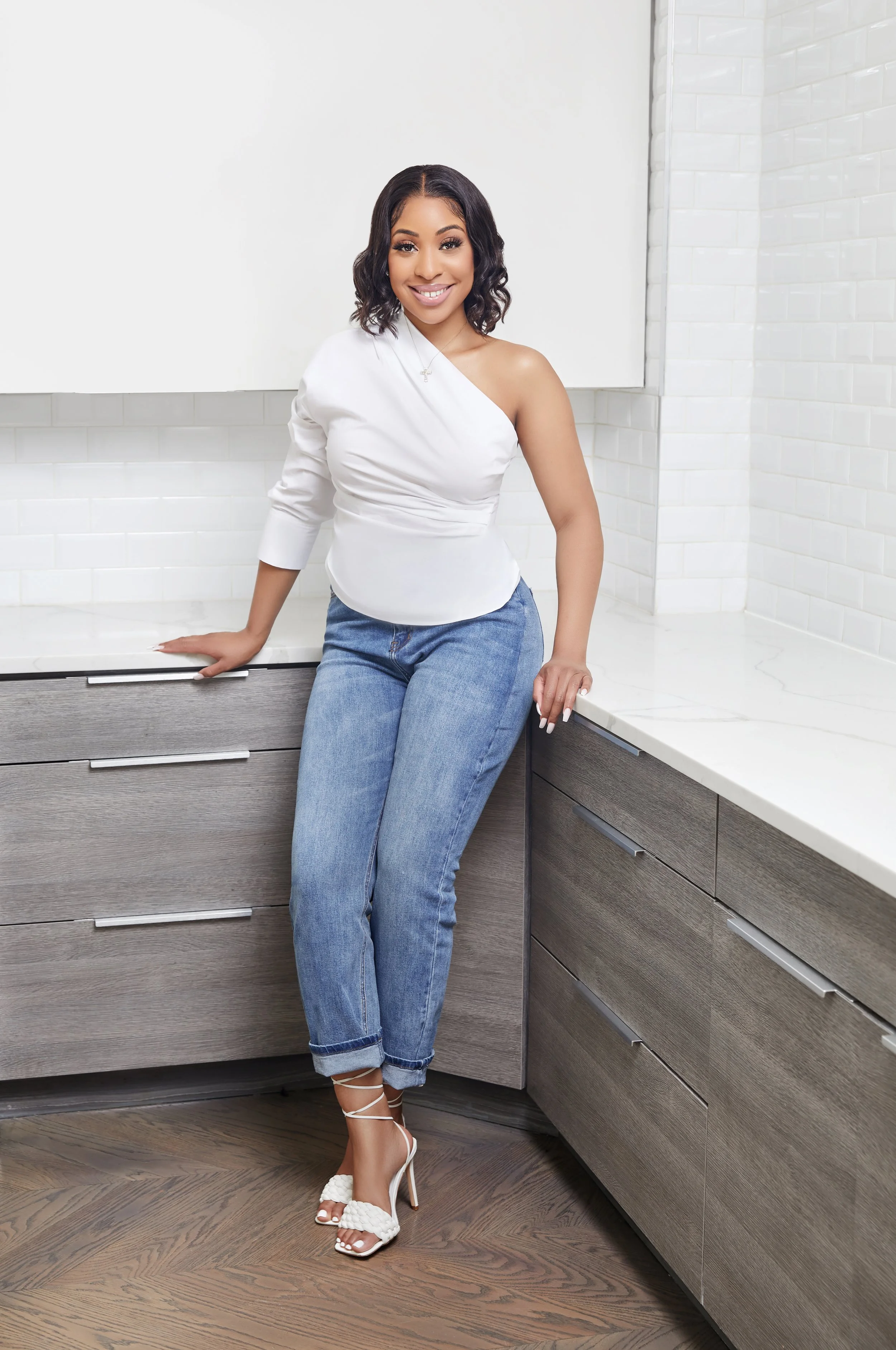 A woman with shoulder-length dark hair, wearing a white asymmetrical top, blue jeans, and white high-heeled sandals, standing in a modern kitchen with wooden cabinets and white tile walls.
