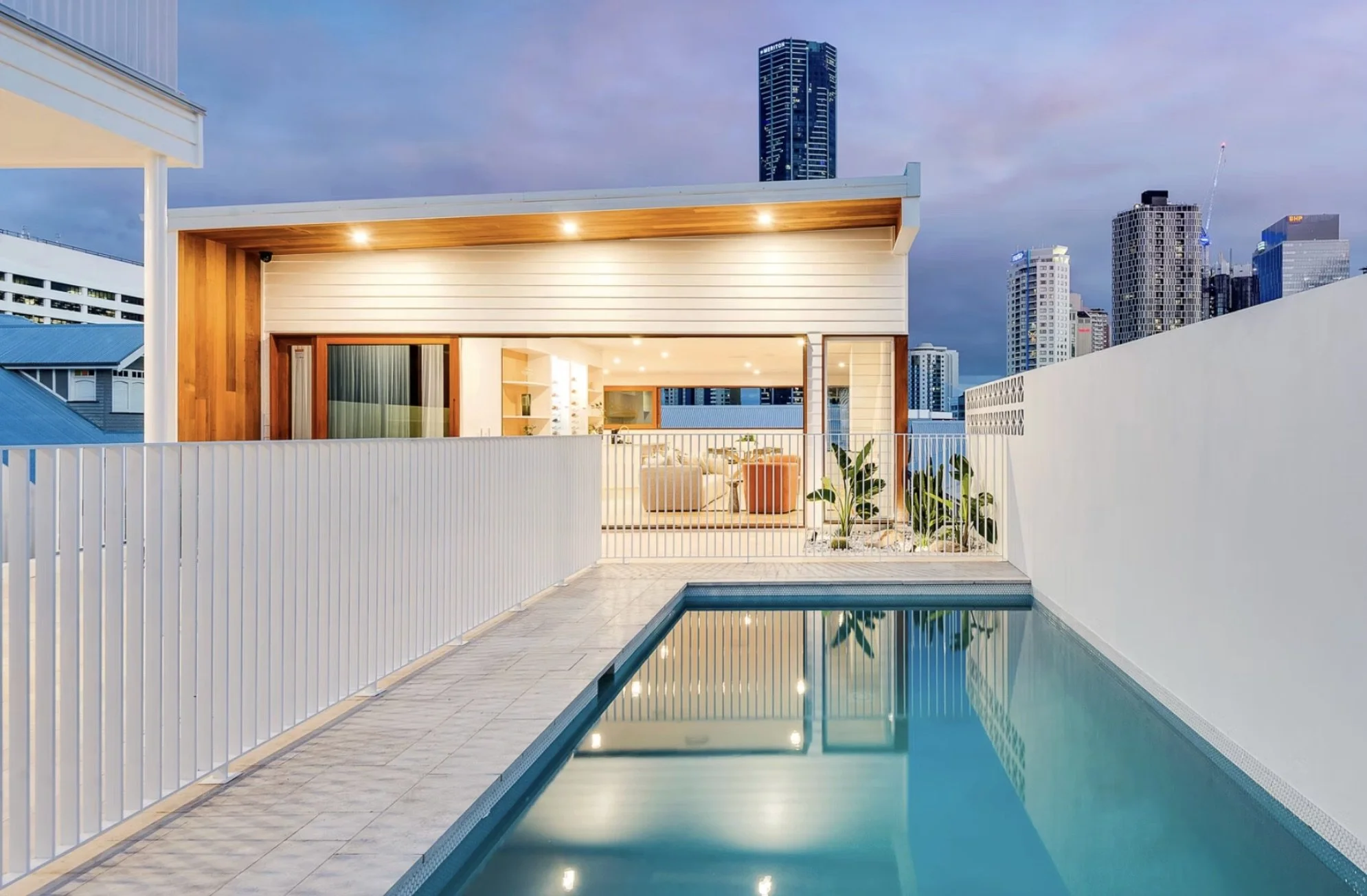 Modern rooftop pool area at dusk, overlooking city skyscrapers, with an outdoor patio and illuminated interior living space.