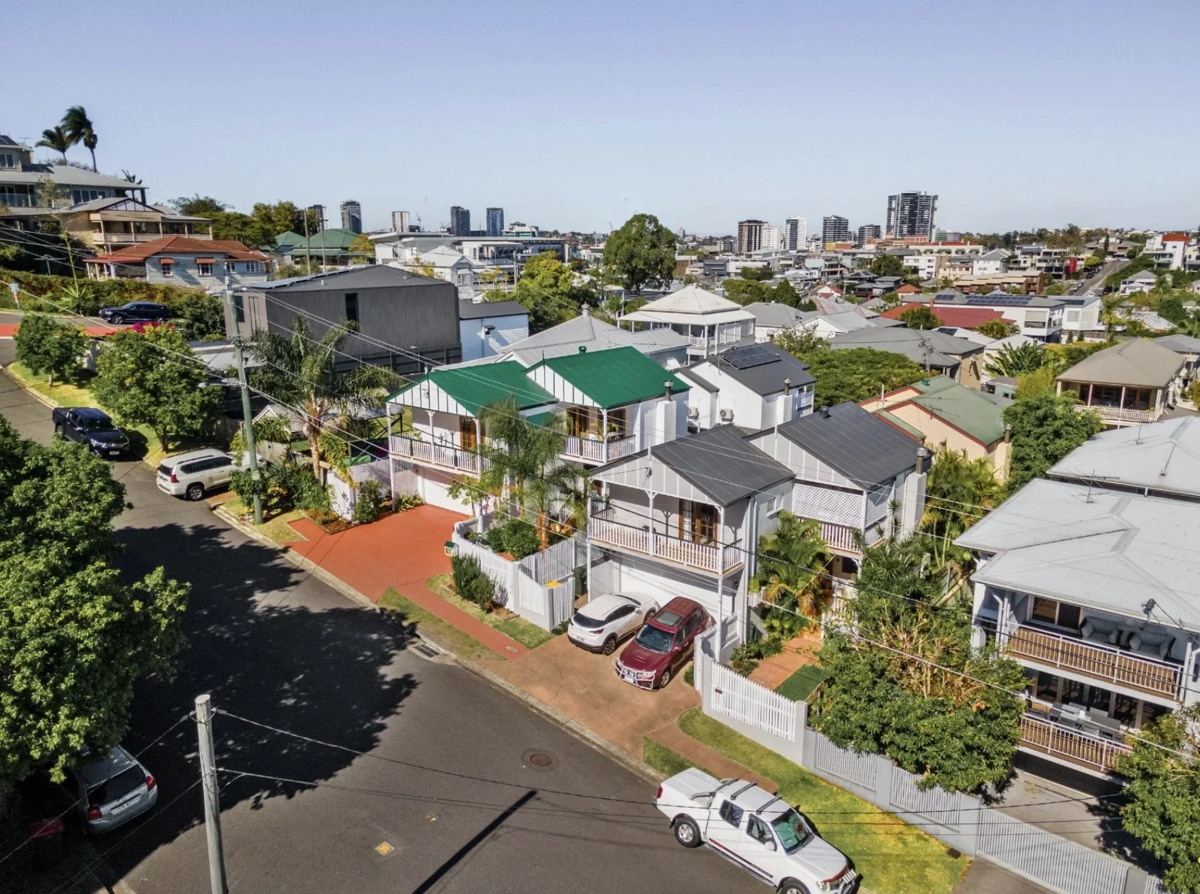 Aerial view of a residential neighborhood with houses, parked cars, trees, and a city skyline in the background.