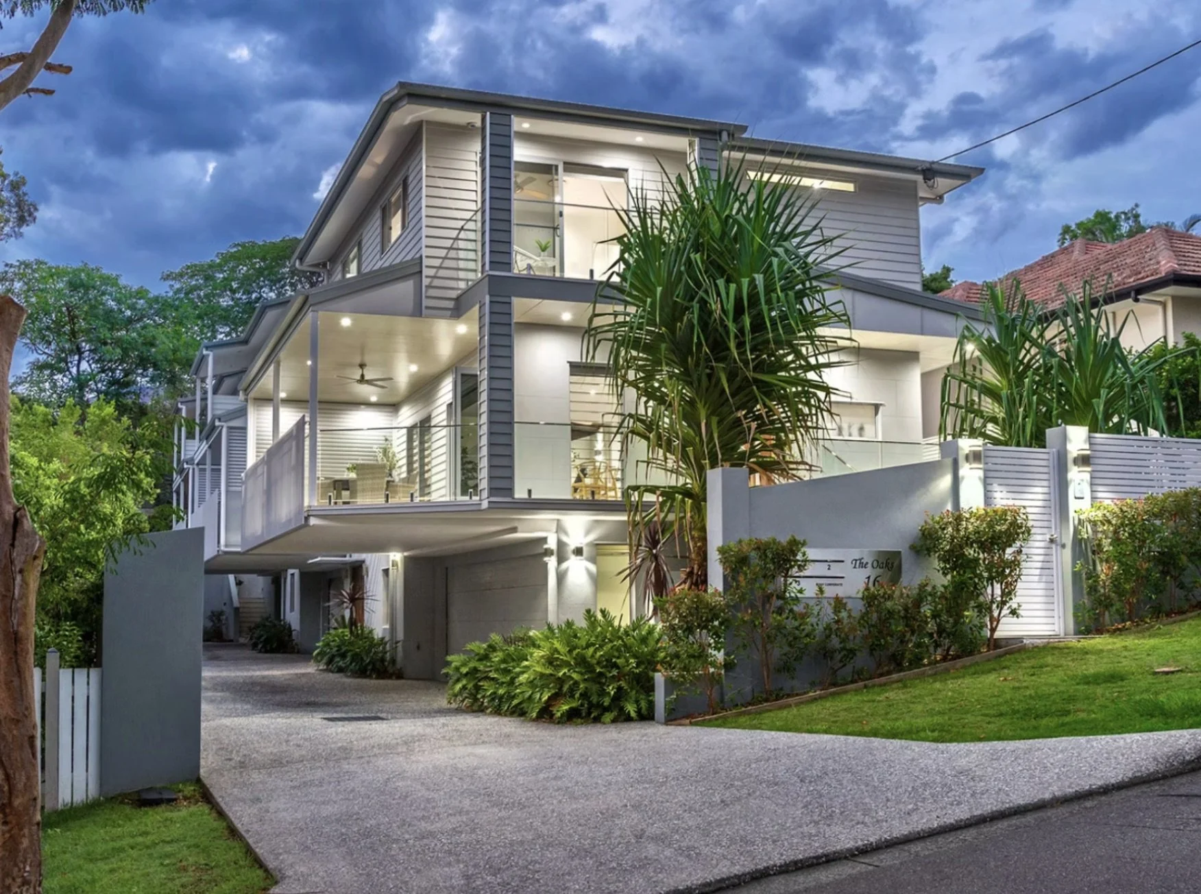 Modern multi-story residential house with large balconies, glass railings, and lush greenery, illuminated with exterior lights at dusk.