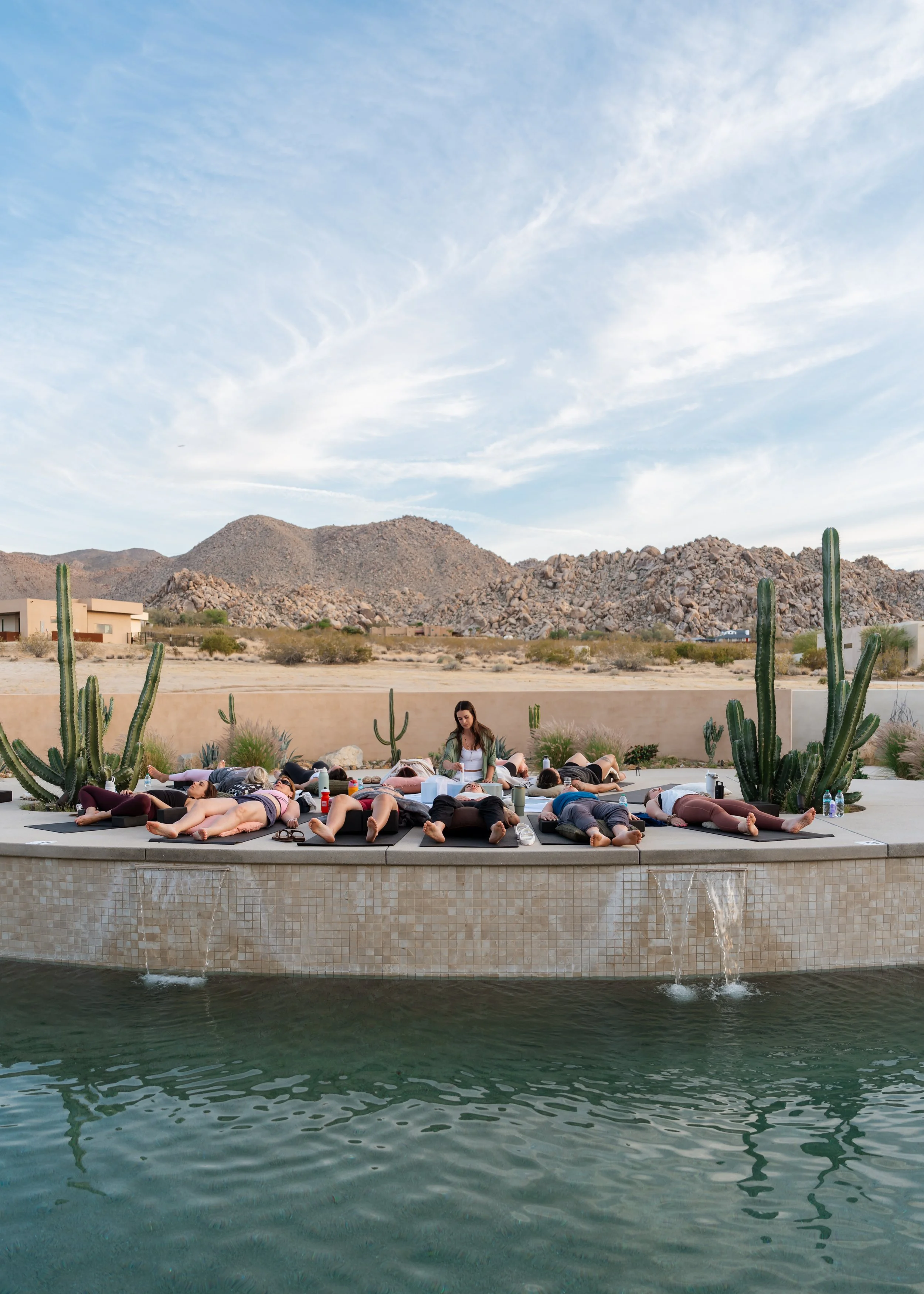 A group of people practicing yoga outdoors on mats around an outdoor pool, with a desert landscape and mountains in the background and cacti in the foreground.