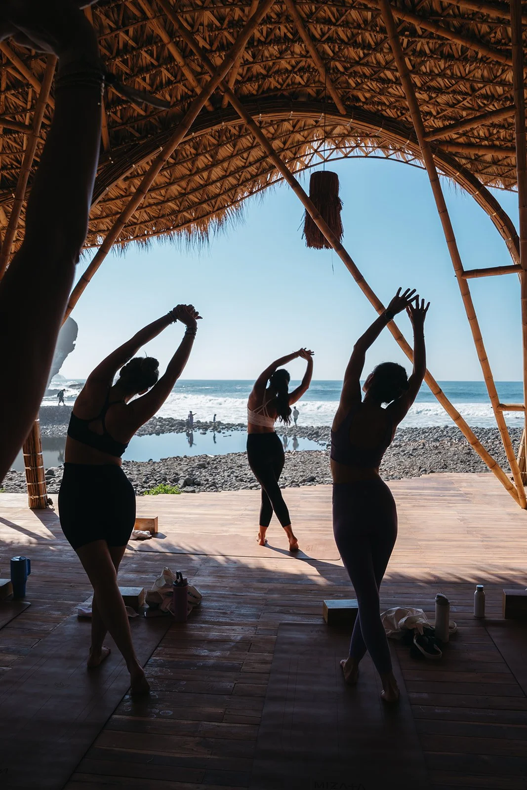 Women practicing yoga on a shaded deck overlooking the ocean with waves and rocks in the background.