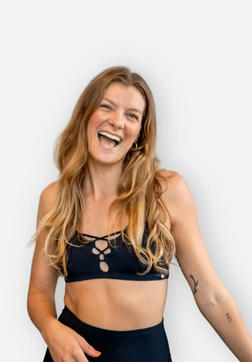 Juliana standing in front of a white wall wearing a black sports bra and leggings with an excited and welcoming expression on her face.