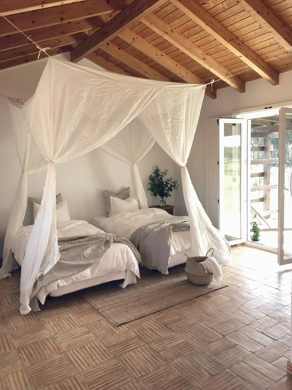 A cozy bedroom with two beds covered in white linens, topped with a canopy made of sheer white fabric. The room has wooden ceilings and floors, a small rug, a potted plant on a bedside table, and an open door leading outside to a wooden deck.