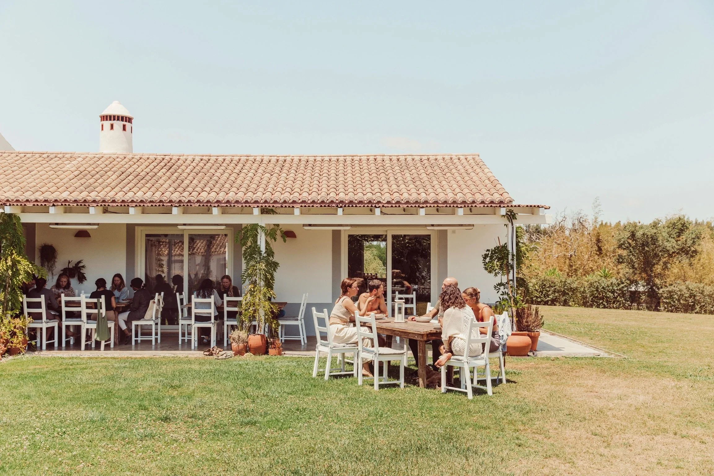 People sitting and talking at white tables with chairs outside a building with a red tile roof, surrounded by greenery and trees.