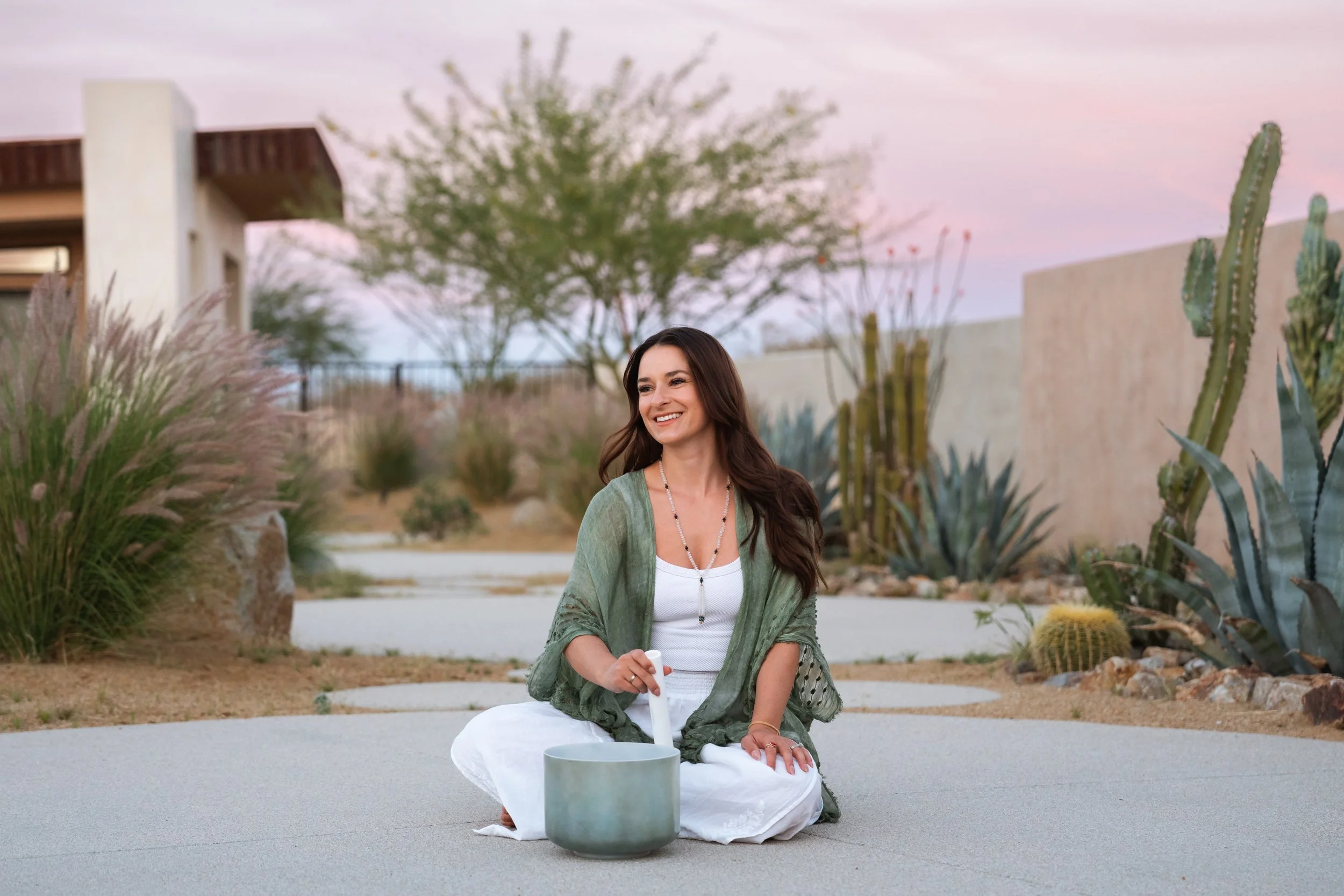 A woman with long brown hair sitting cross-legged on the ground outdoors, smiling, with a cactus and desert plants in the background, and a pastel-colored sky.
