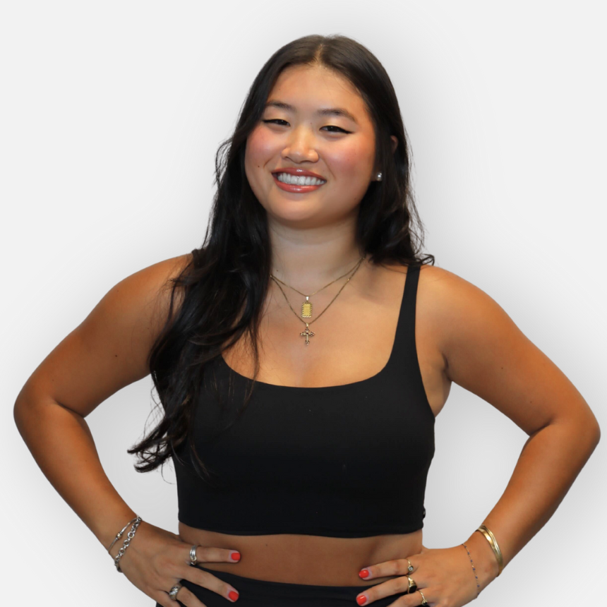A smiling woman with long black hair wearing a black tank top, gold jewelry, and standing with her hands on her hips.