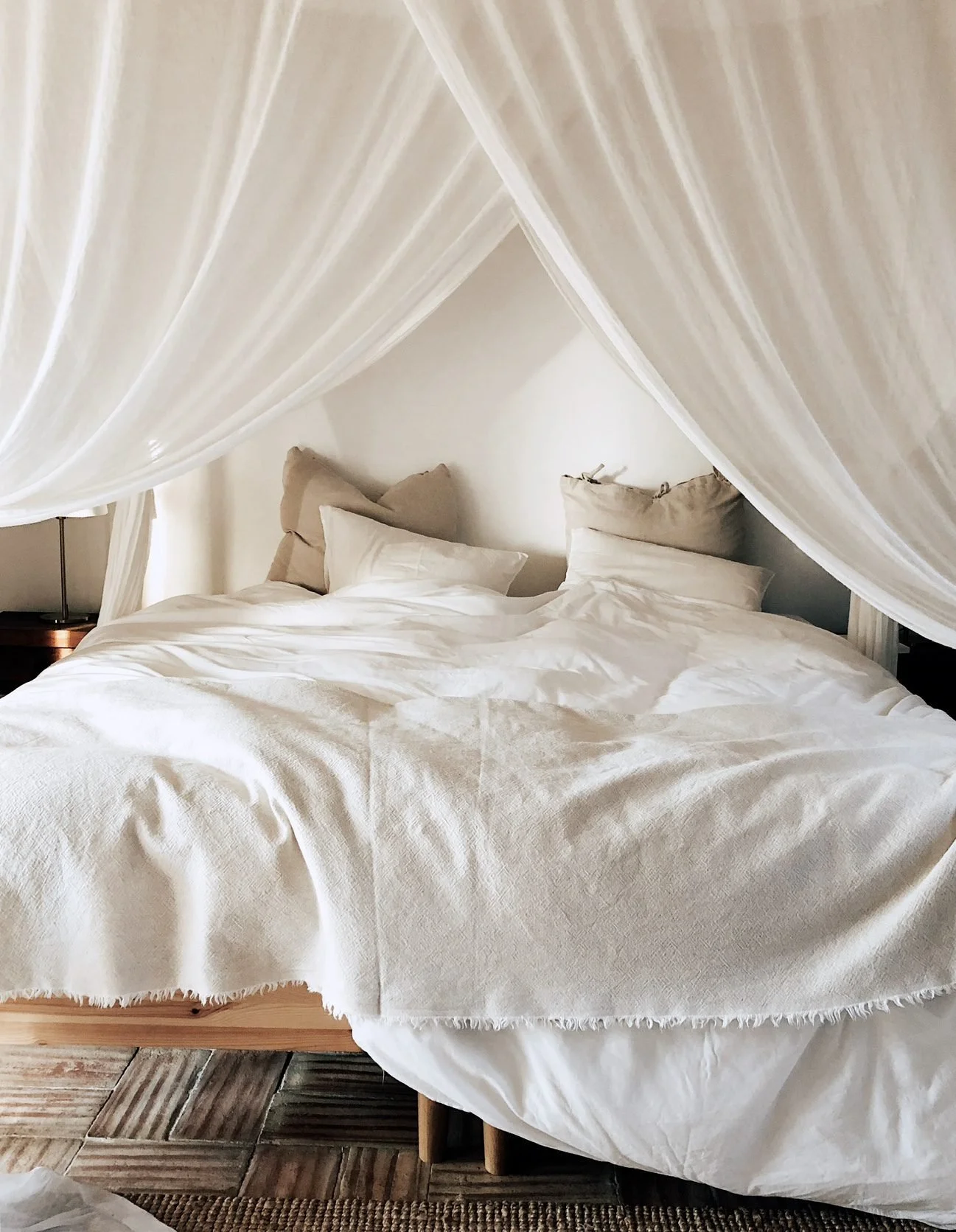A bed with white bedding and beige pillows, surrounded by a white canopy or drapes hanging from above.
