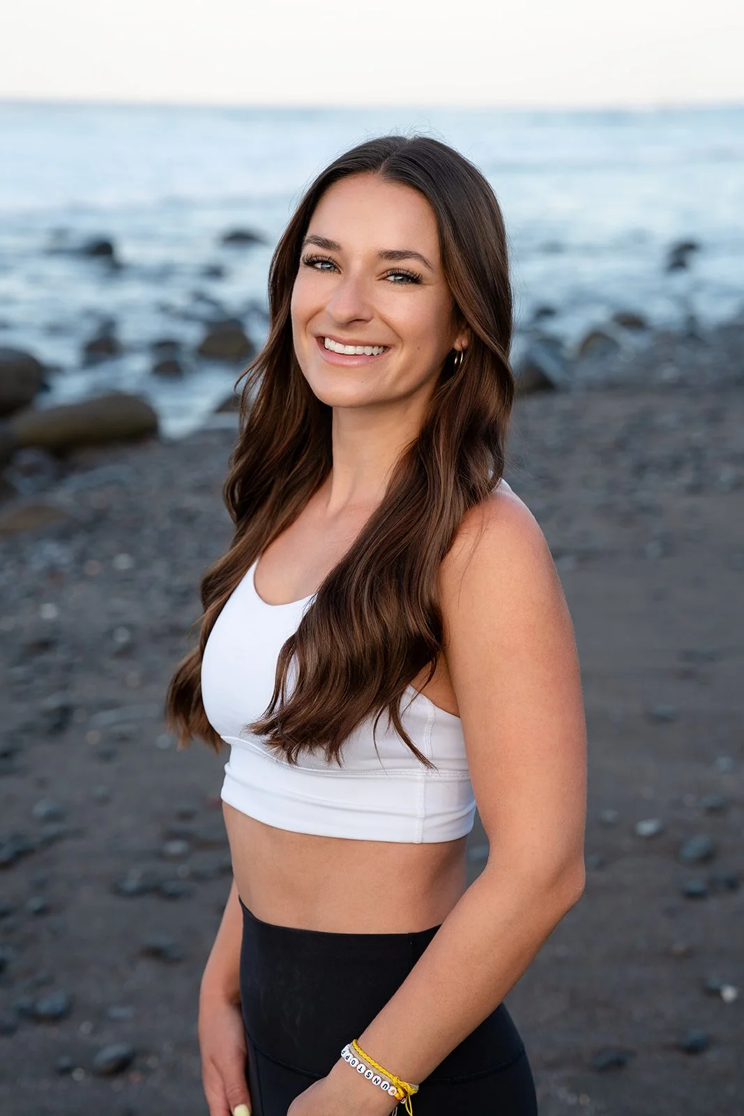 A woman with long wavy brown hair smiling at the camera on a rocky beach with water in the background.
