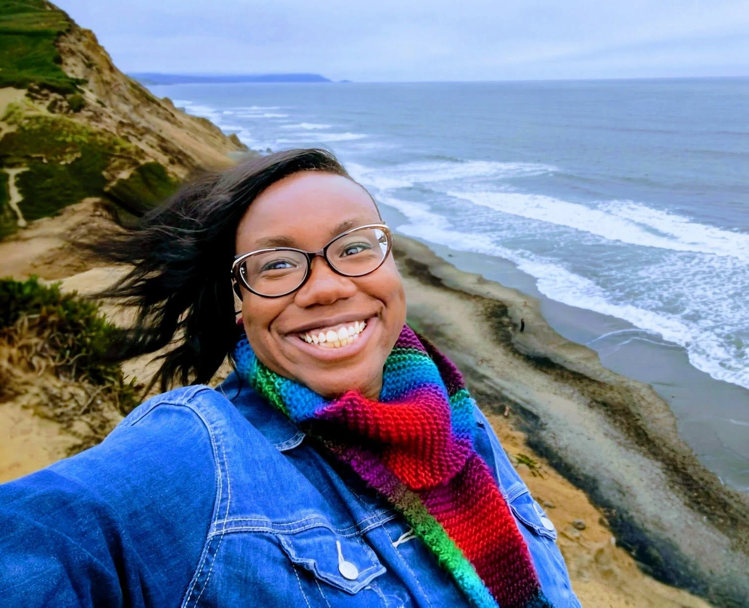 A smiling woman with glasses and a colorful scarf taking a selfie at the edge of a cliff overlooking the ocean, with waves crashing on the beach below and green hills in the background.