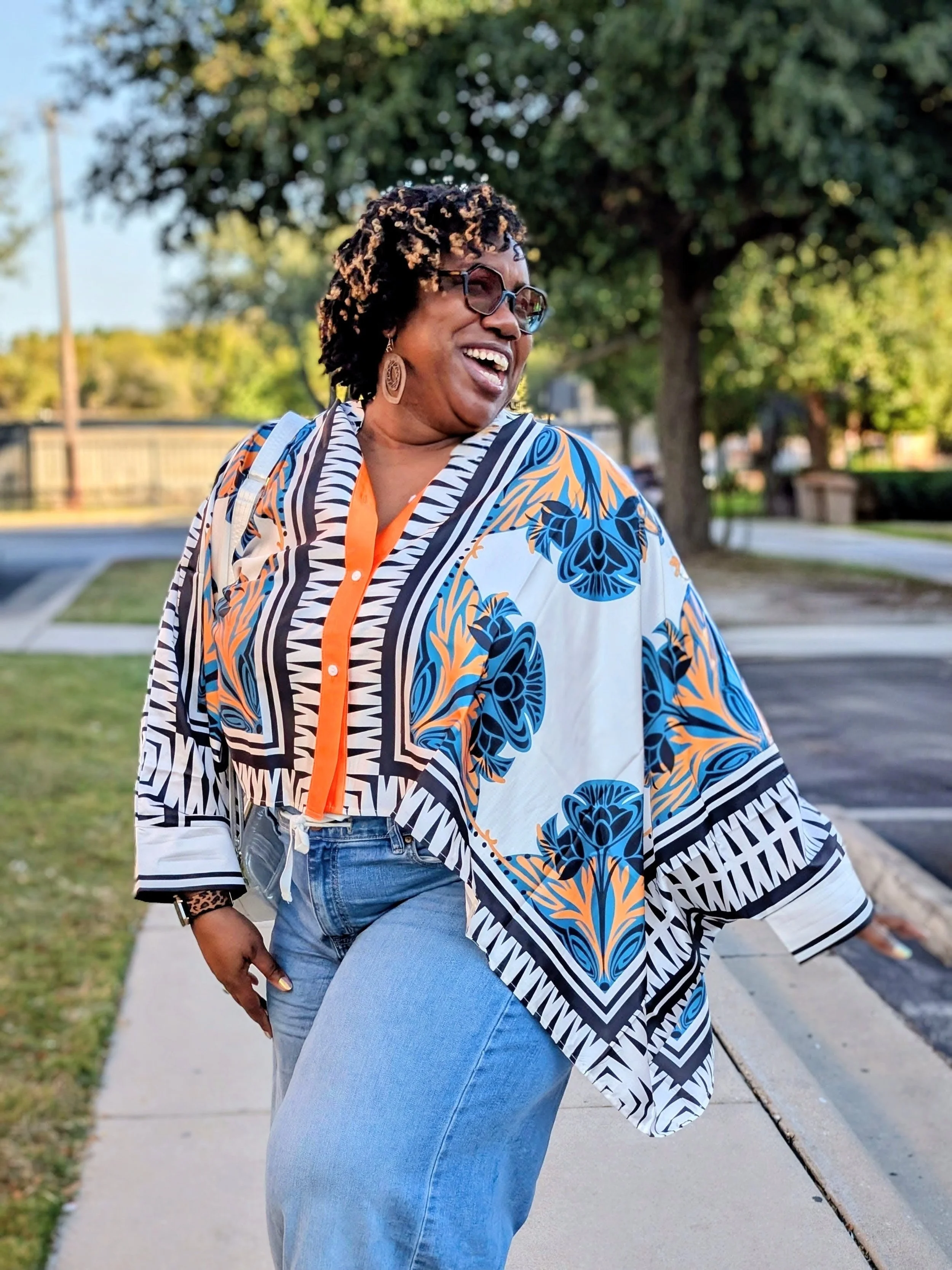 A joyful woman with short curly hair, wearing glasses, an ornate black, white, blue, and orange patterned blouse, blue jeans, and earrings, laughing outdoors in a park.
