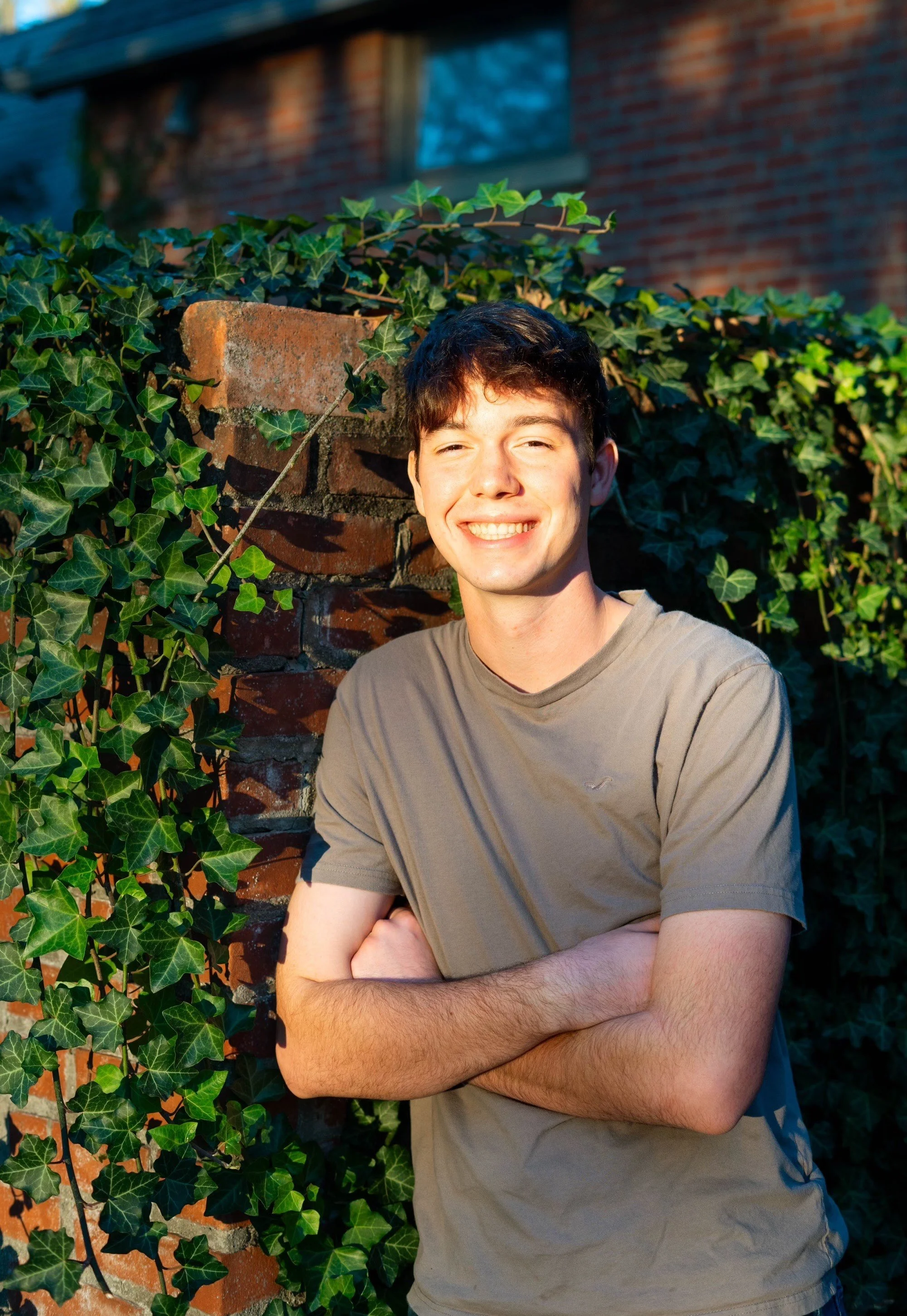 Young man smiling outdoors, standing with arms crossed in front of a brick wall and green ivy plants.