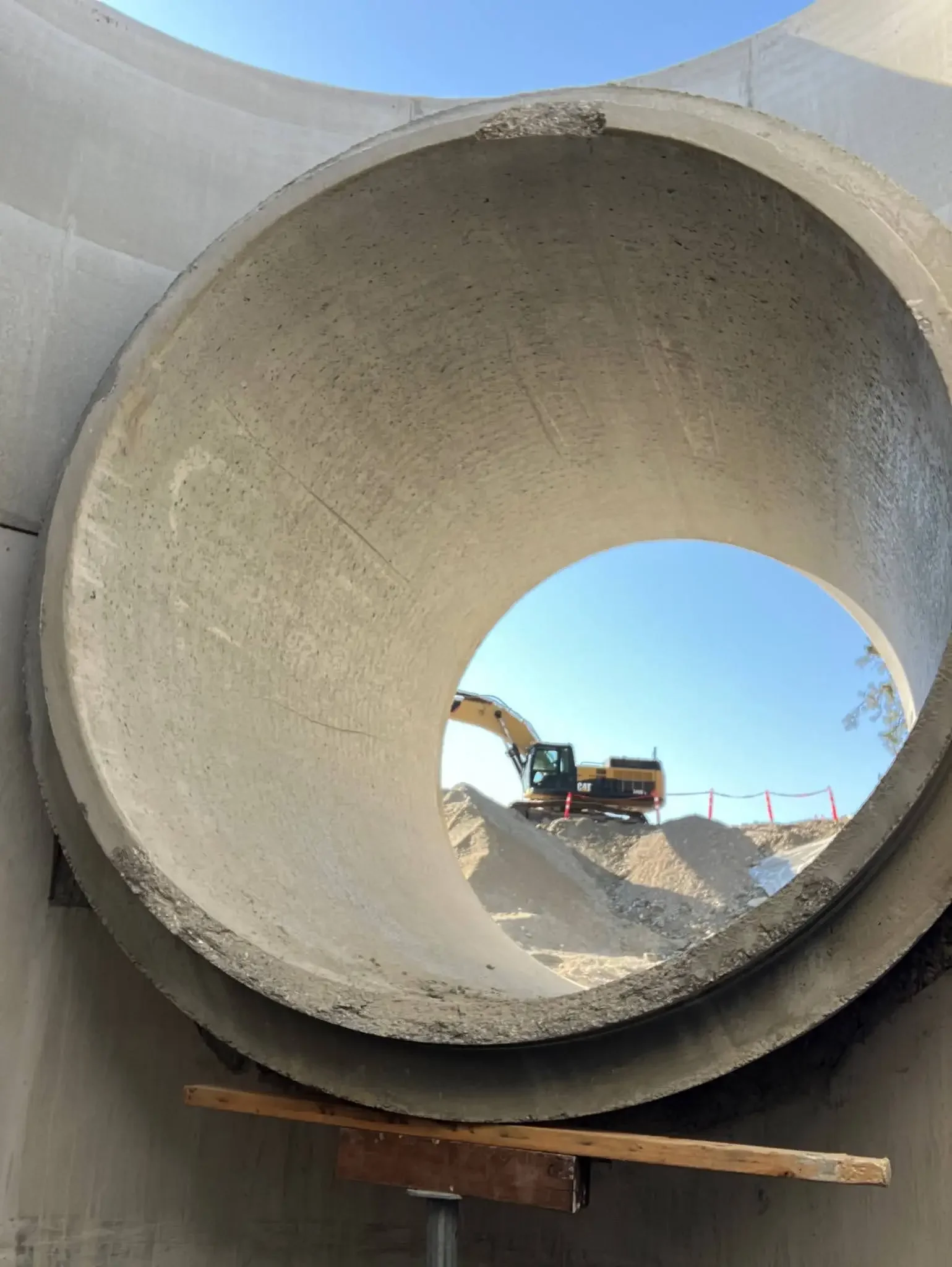Large concrete pipe with a construction excavator visible through the opening, set on a construction site under a clear blue sky.