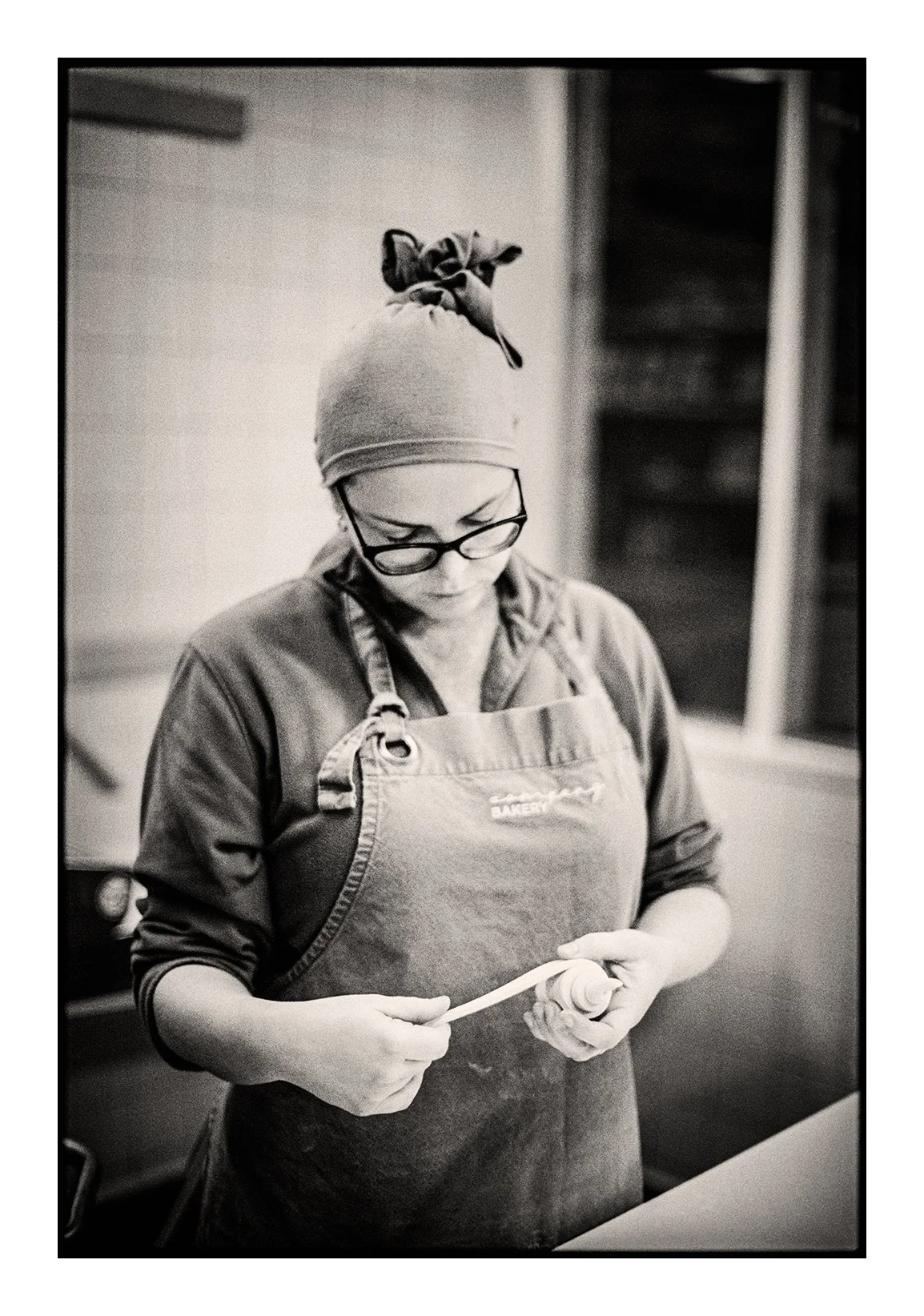 A Photograph by Colin I Homes of a  woman wearing glasses, a head scarf, and an apron, looking down and making a croissant.