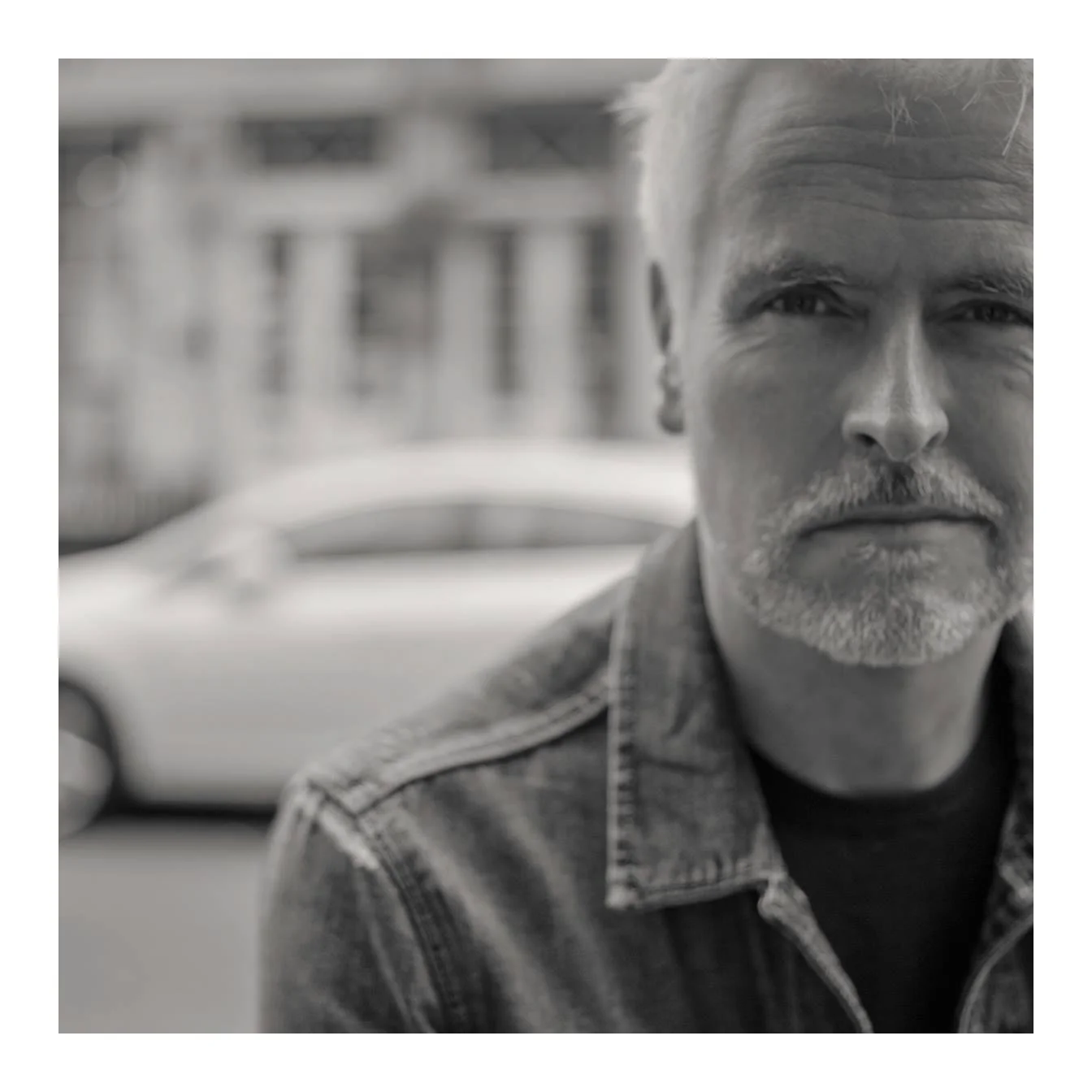 A close-up black and white portrait of Colin Ironside Homes, wearing a denim jacket, with buildings and a car in the background.