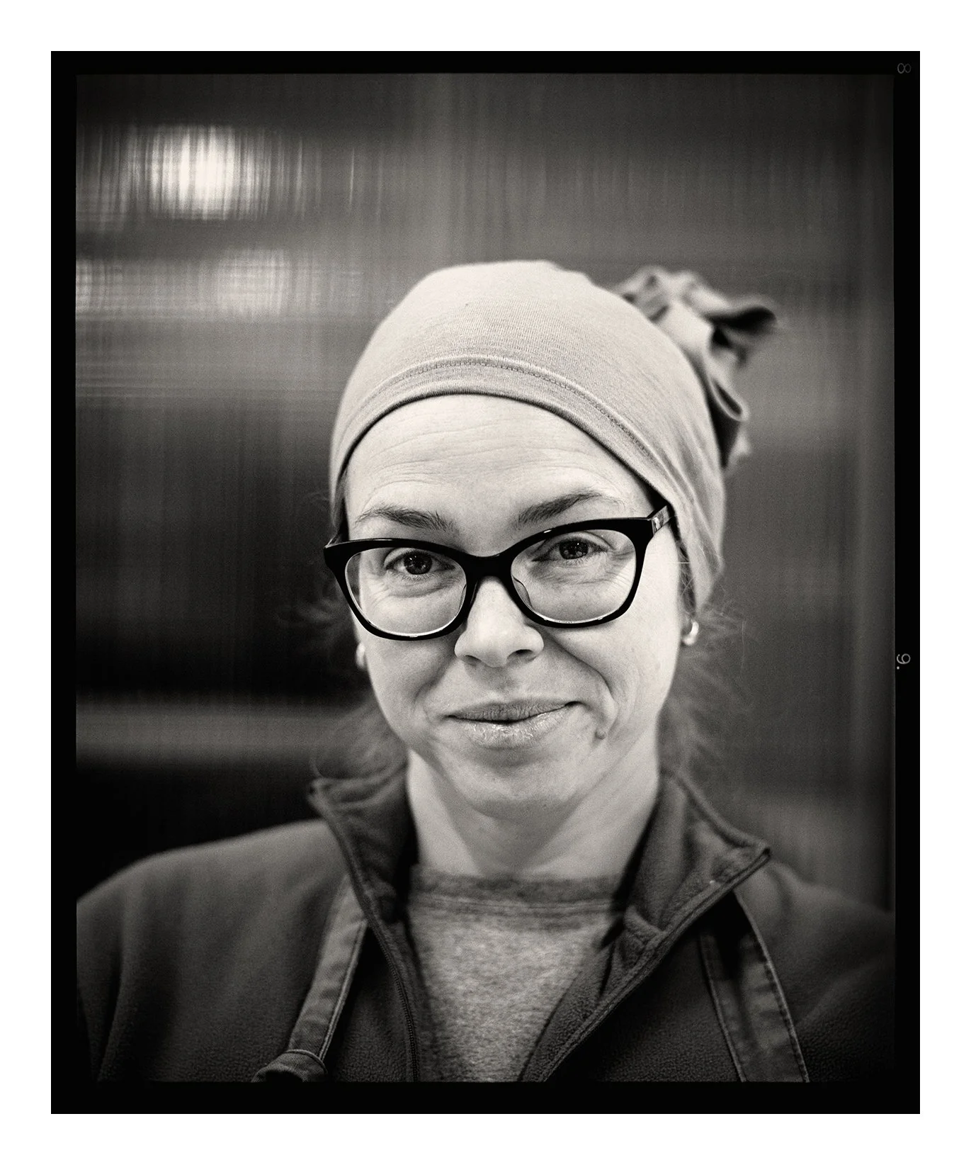 Black and white portrait by fine art portrait photographer Colin I Homes of a woman wearing glasses, a headscarf, and a jacket, smiling at the camera.