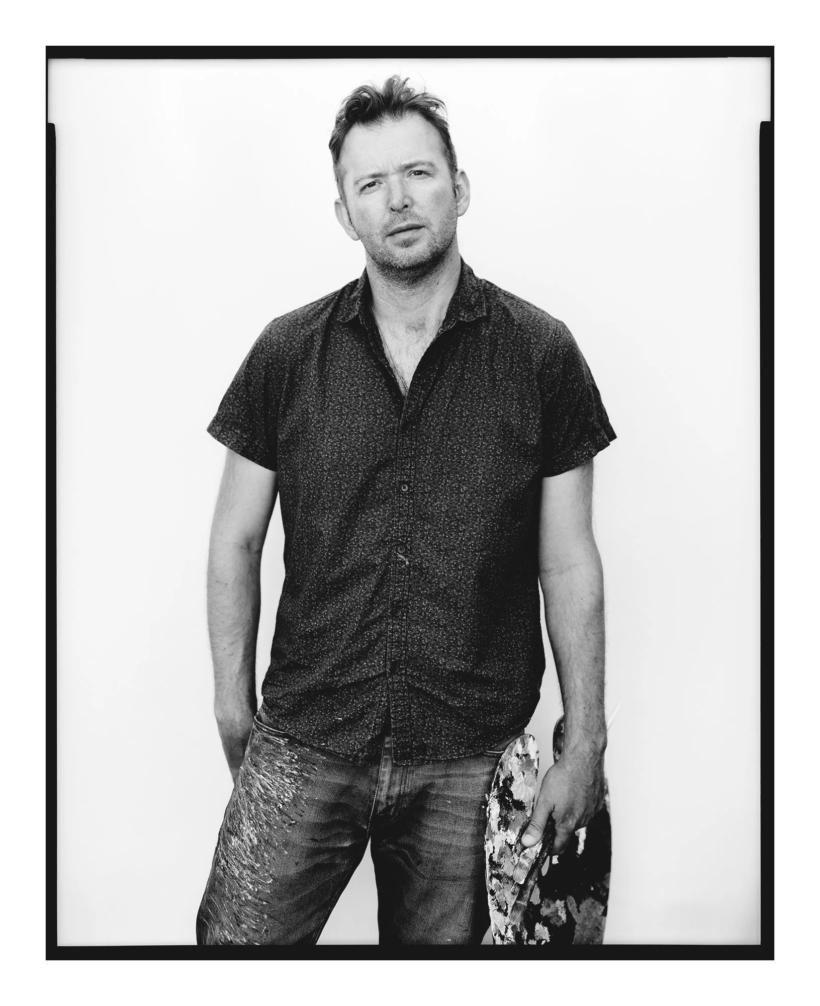 Black and white portrait of a man with short hair, wearing a button-up shirt, holding a paint palette and brush, standing against a plain background.
