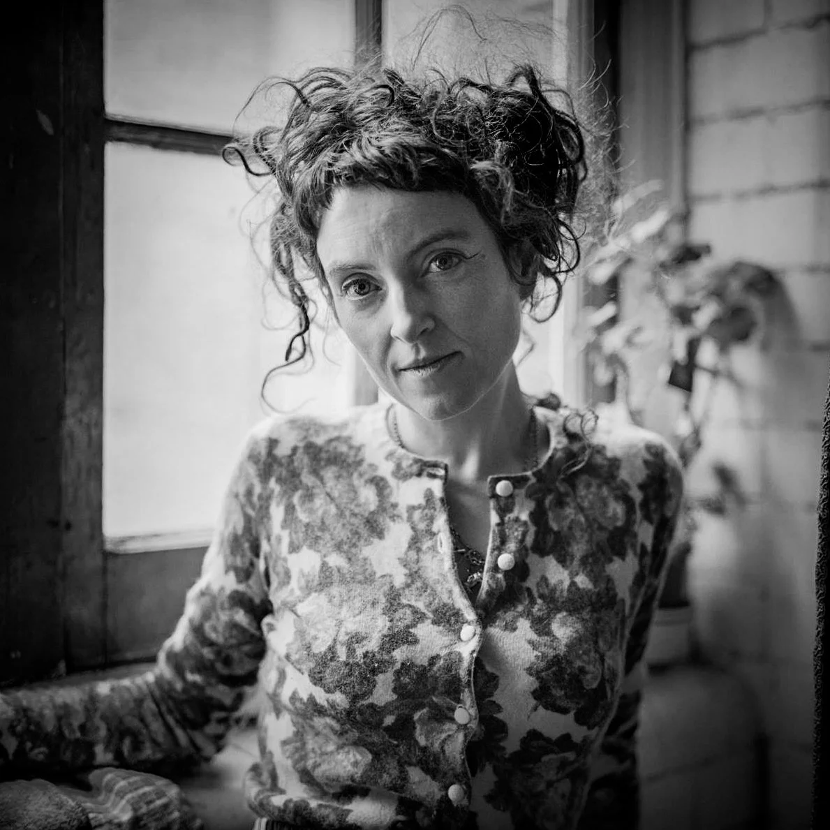 A black and white portrait by fine art photographer Colin I Homes of a woman with curly hair, wearing a floral-patterned top, looking at the camera with a gentle expression, positioned near a window with light illuminating her face.