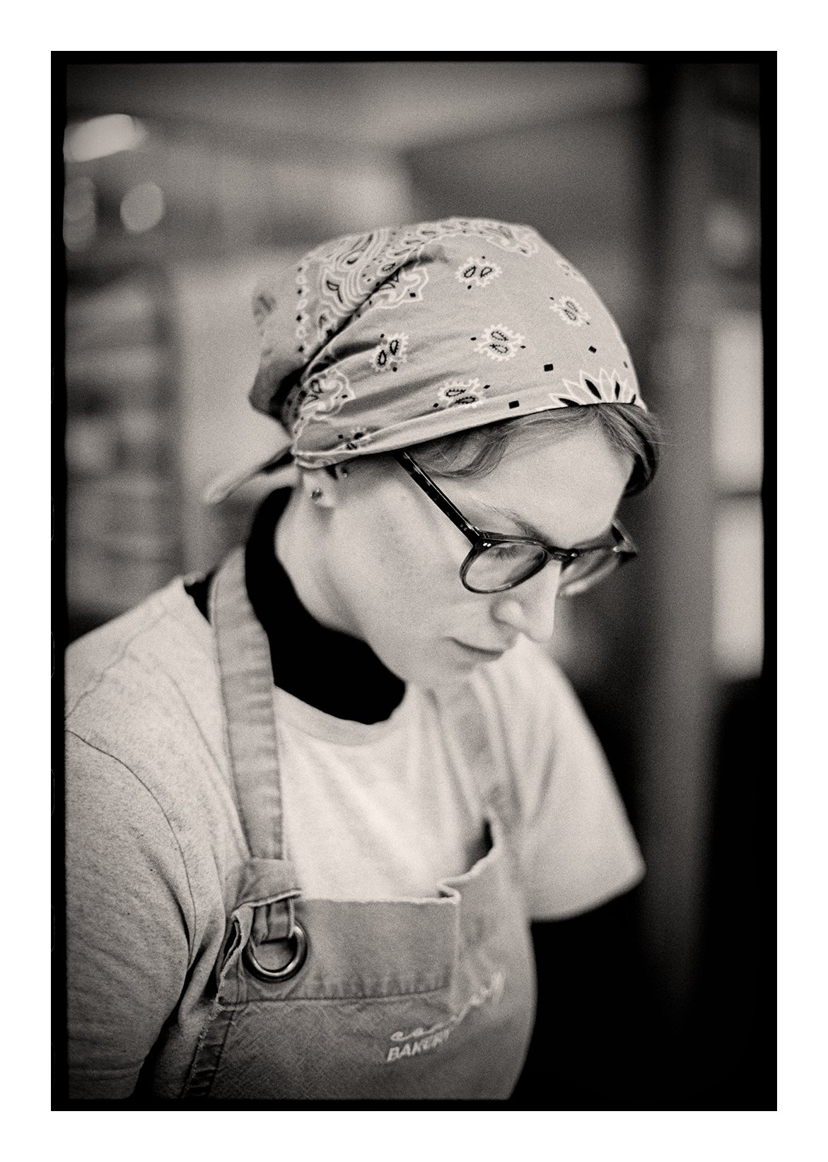 A photograph by Colin I Homes of a woman wearing glasses, a bandana, and an apron, focused on baking  in a kitchen.