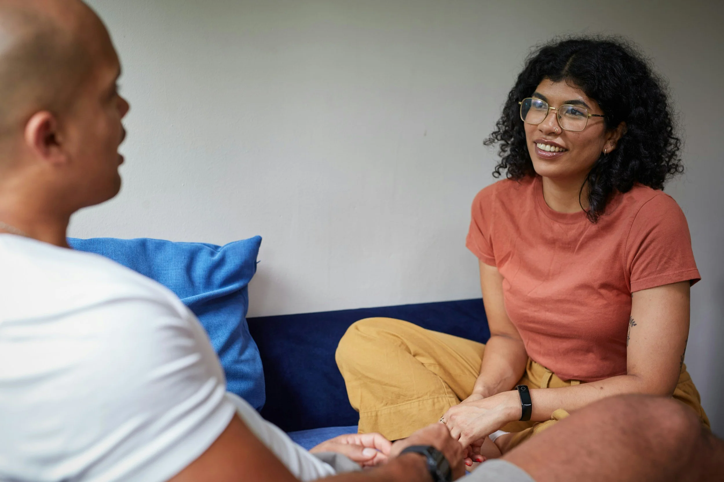 A woman with curly black hair, glasses, and a nose ring, smiling and talking to a man with a shaved head, sitting on a blue couch, holding hands.