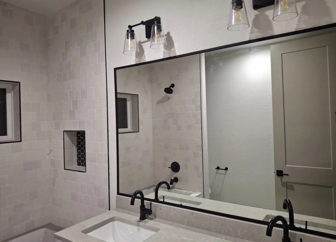 A modern bathroom with dual black sinks, a large mirror, and a wall-mounted lighting fixture with three glass shades. The bathroom features beige tiled walls, a shower with a black showerhead, and small windows with black trim.
