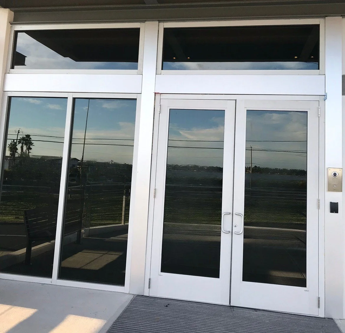 Exterior glass doors of a modern building with reflective windows showing an outdoor landscape, including trees, power lines, and a cloudy sky.