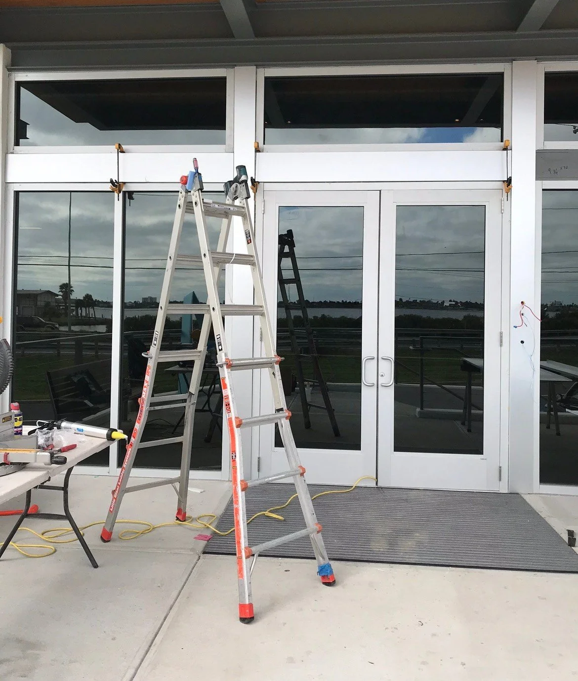 Ladder and tools in front of a building with glass doors and windows, under an overcast sky.