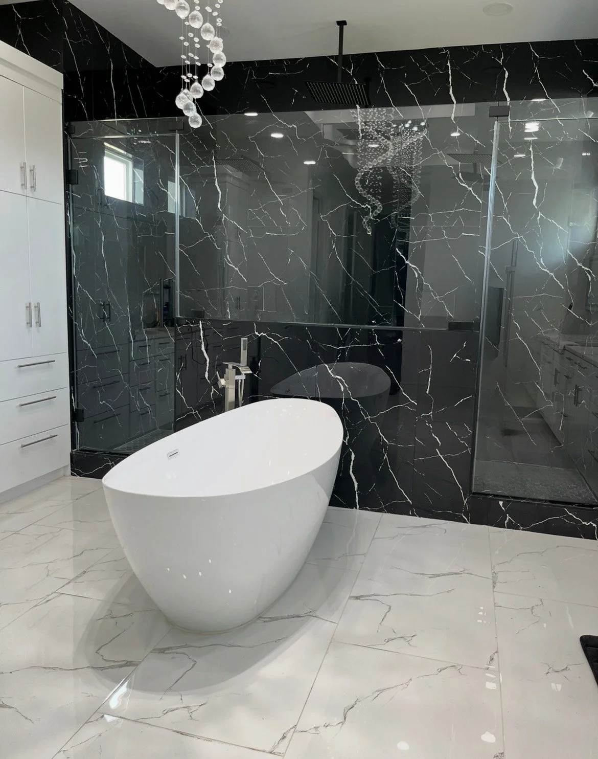 Modern bathroom with a freestanding white bathtub, black marble wall, glass shower enclosure, white cabinetry, and marble flooring.