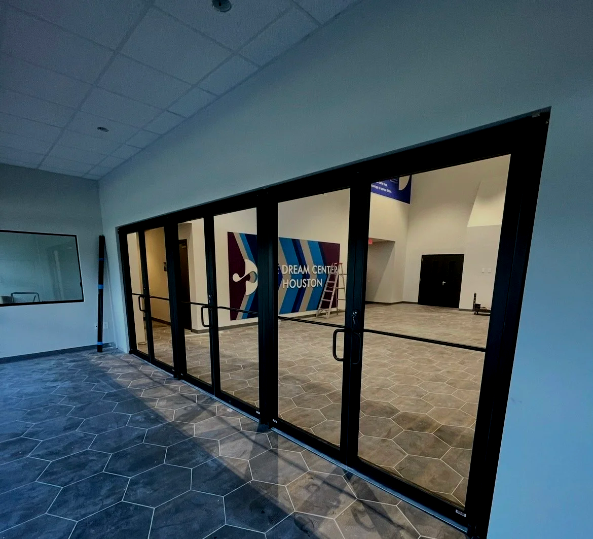 Interior view of the Houston Dream Center, showing a glass entrance with doors, hexagonal tile flooring, and a mural on the wall inside featuring a face with the text 'DREAM CENTER HOUSTON'.